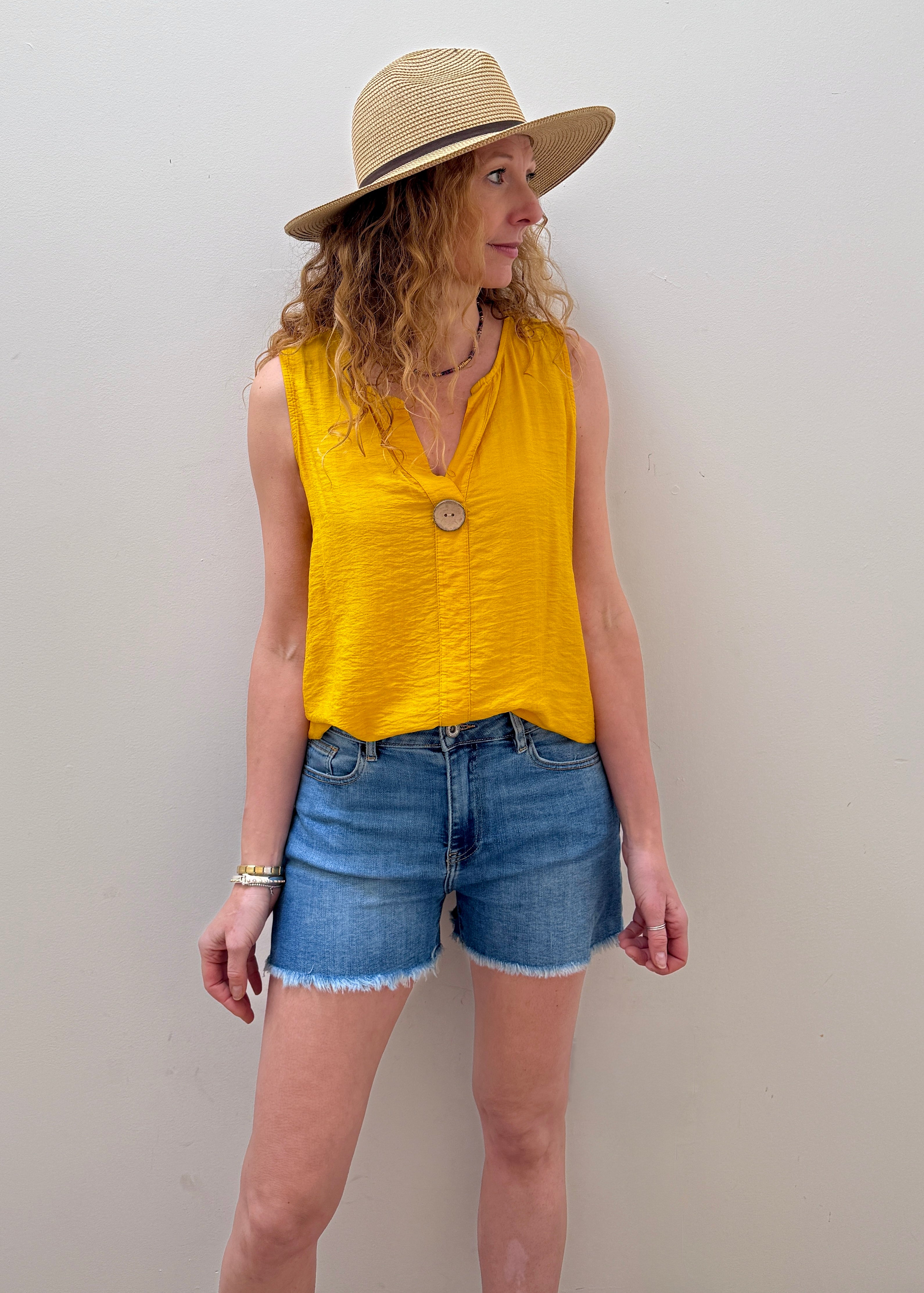 Woman wearing a yellow sleeveless top and denim shorts with a straw hat against a plain background