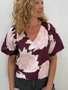 A woman with curly blonde hair wearing a burgundy and pink floral print blouse with puff sleeves, paired with dark gray pants, standing against a plain white wall.