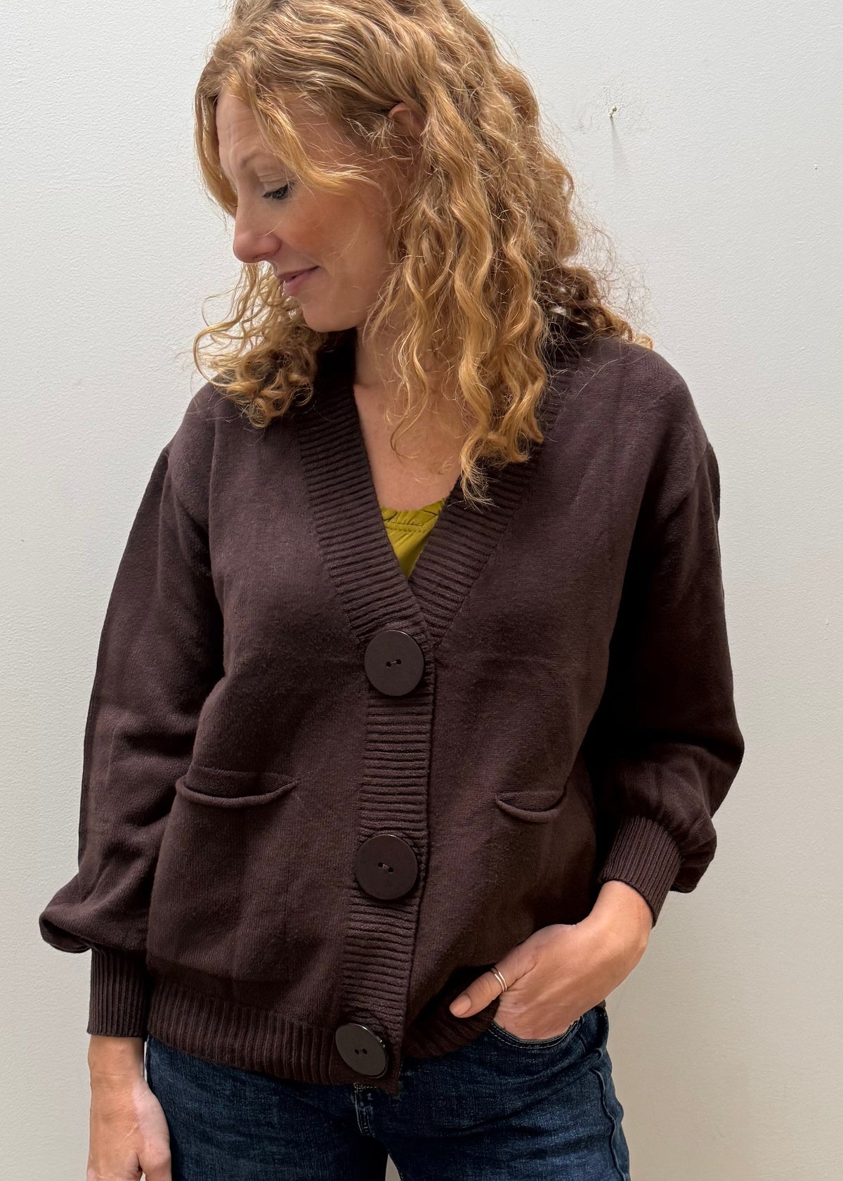 A woman wearing a dark brown button-front cardigan and blue jeans, standing in a room with a plain white wall.