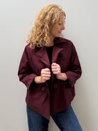A woman with curly blonde hair wearing a dark burgundy button-up jacket and blue jeans, standing against a plain light gray wall.