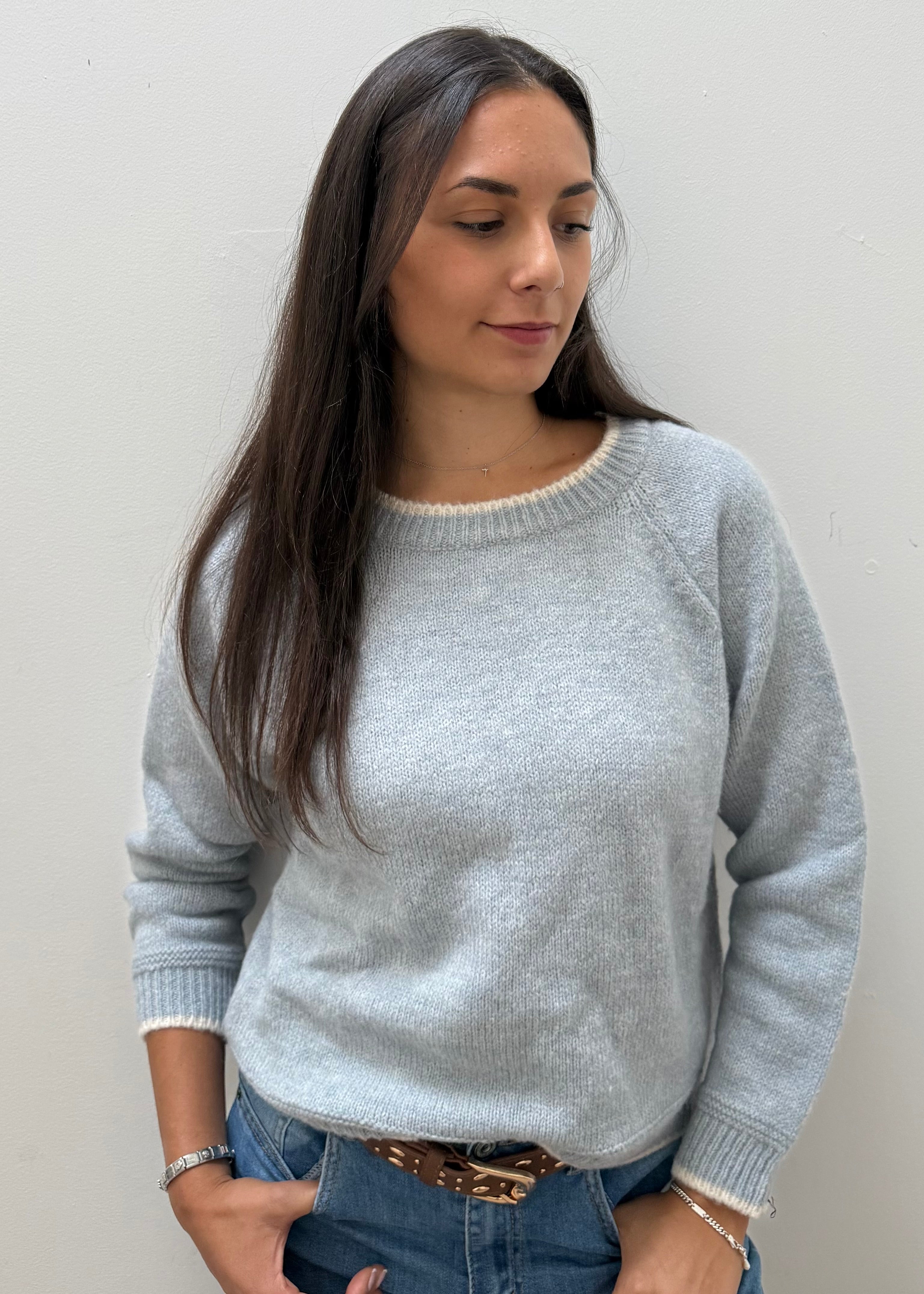 A woman wearing a light gray knit crewneck sweater, paired with blue jeans and a brown belt, standing against a plain light gray wall.