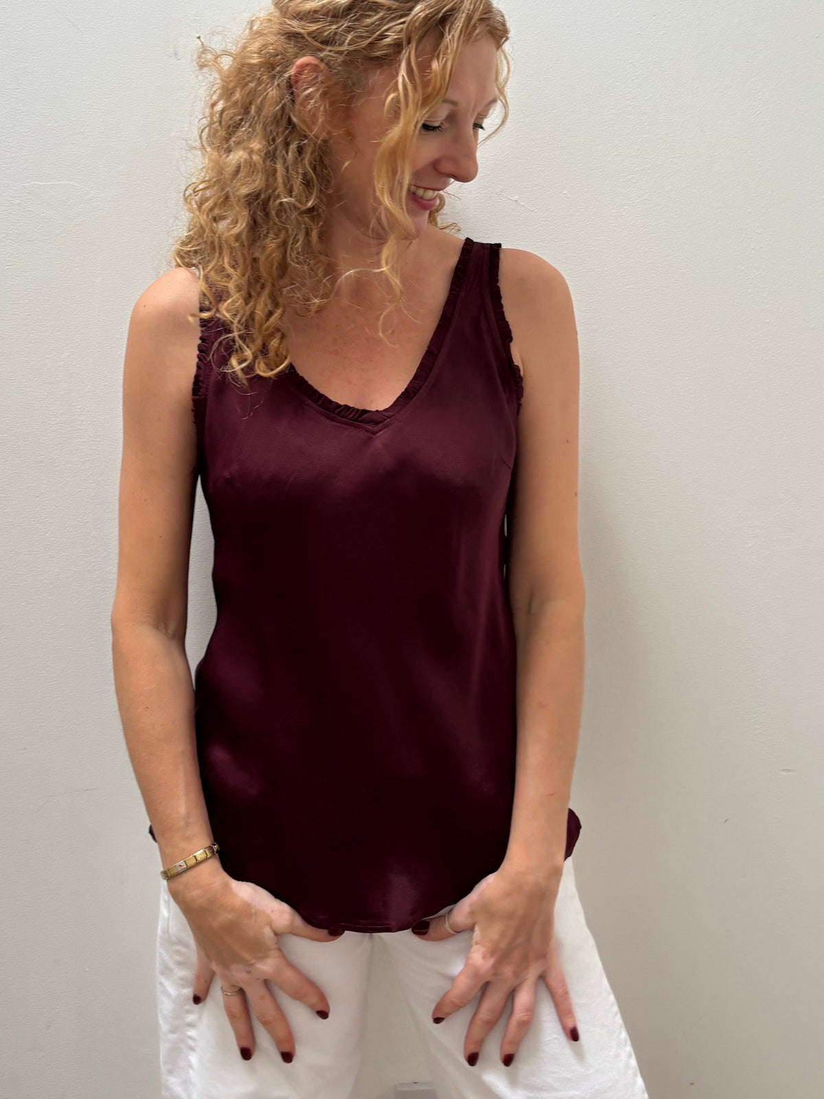 A woman with curly blonde hair wearing a sleeveless, satin-like burgundy tank top and white pants, standing against a plain off-white wall.