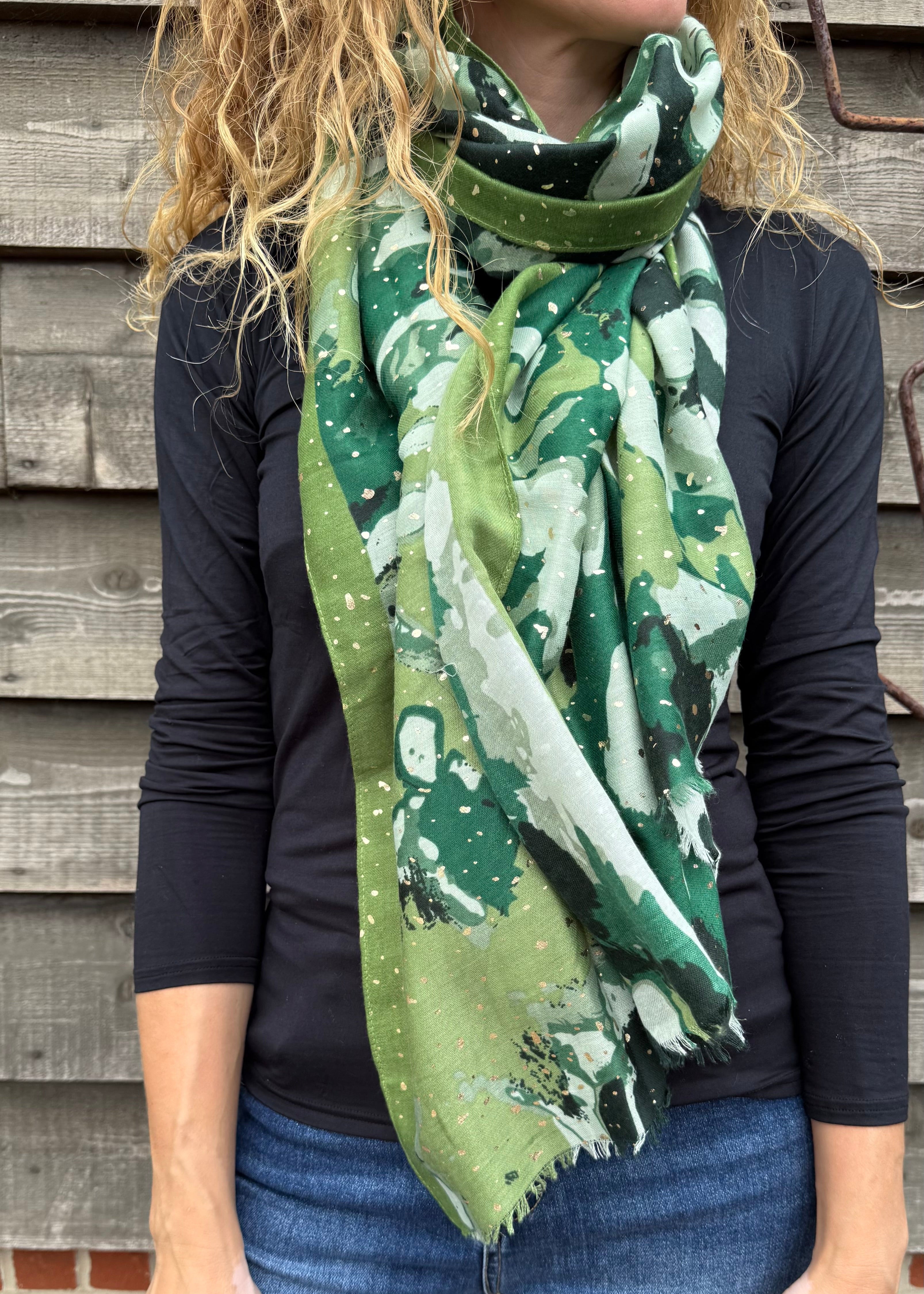 Person wearing a green patterned scarf with a dark shirt and jeans against a wooden wall.