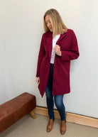 Woman wearing a burgundy coat, white shirt, blue jeans, and brown boots against a plain wall.