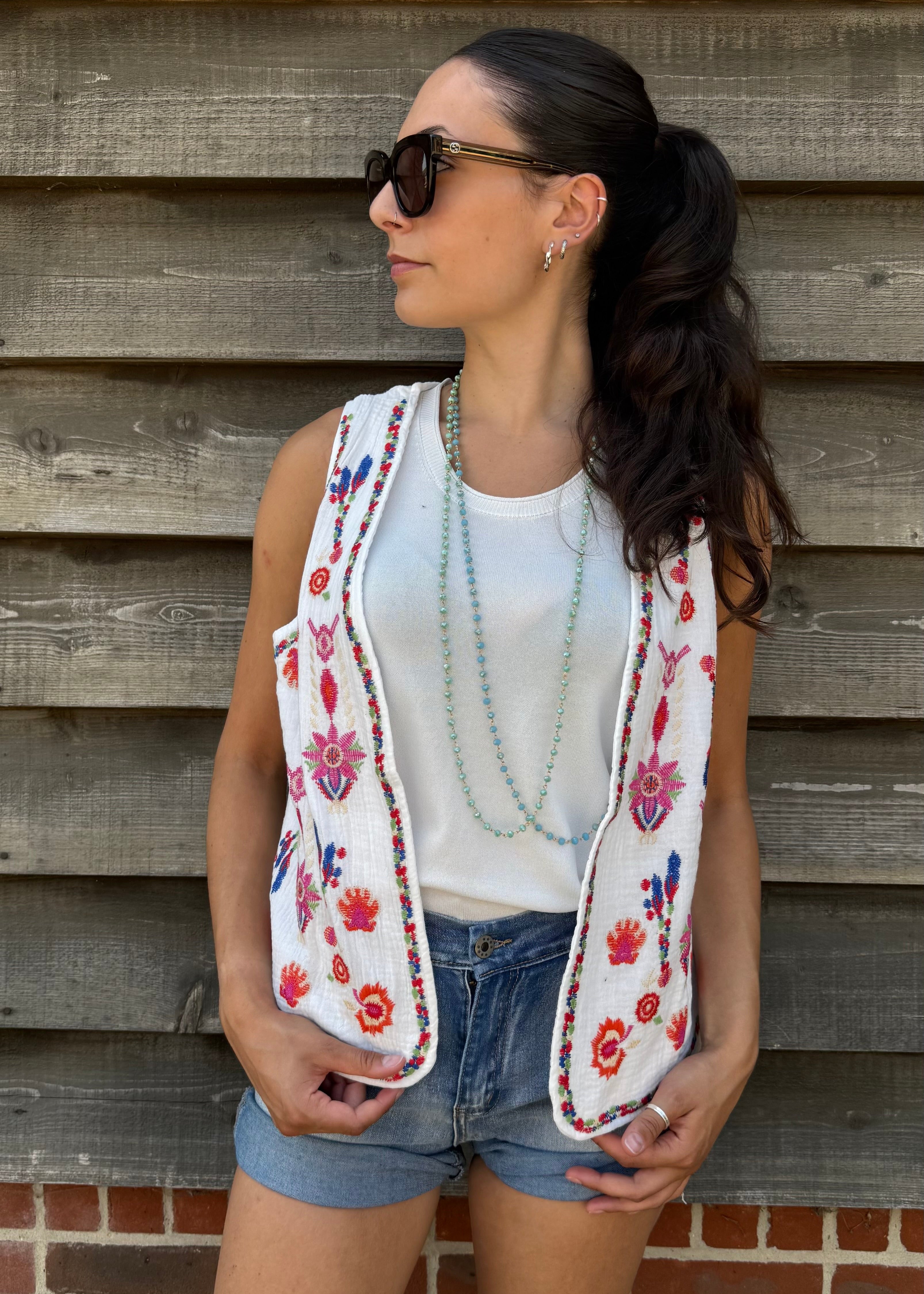 Woman wearing a floral embroidered vest over a white top and denim shorts, standing against a wooden wall.