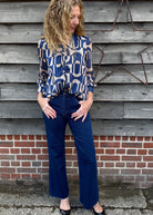 Woman wearing a patterned blouse and blue jeans standing against a wooden wall.