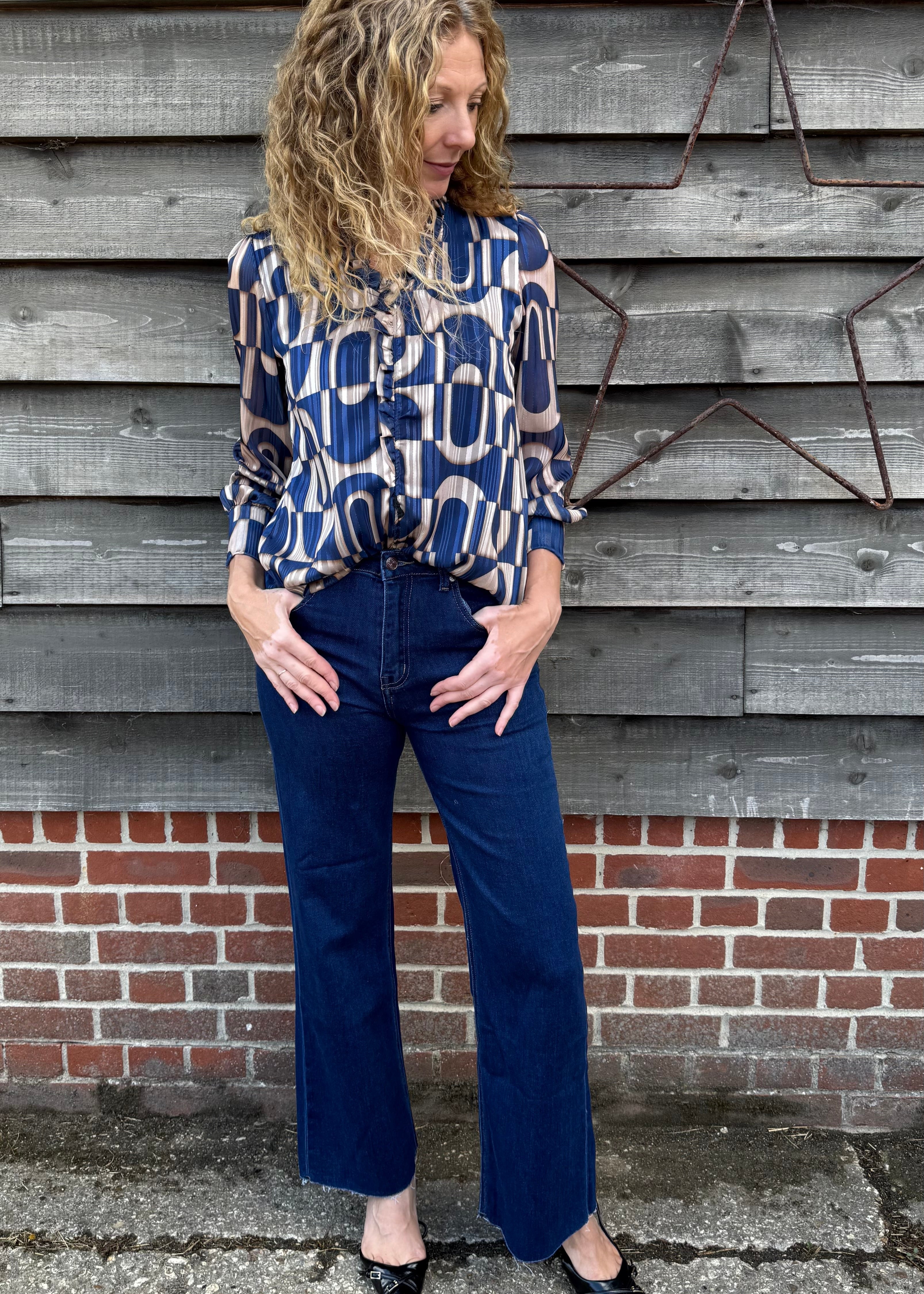 Woman wearing a patterned blouse and blue jeans standing against a wooden wall.