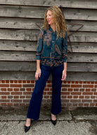 Woman in a patterned shirt and blue jeans standing against a wooden wall.