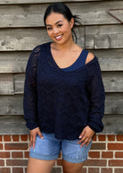 Woman wearing a navy blue sweater with cut-out shoulders against a wooden wall.