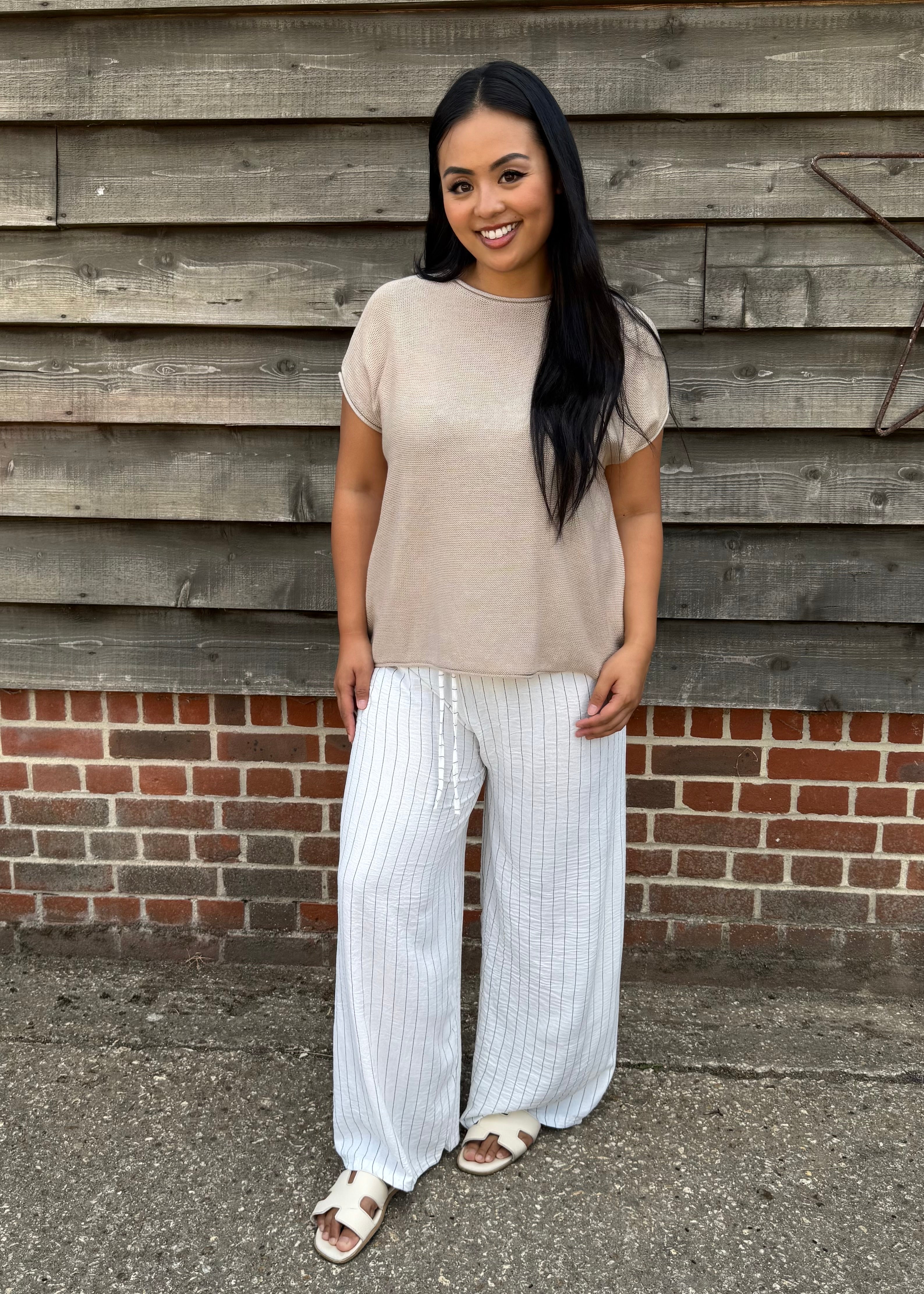 Woman wearing a beige top and white striped pants standing against a wooden wall.