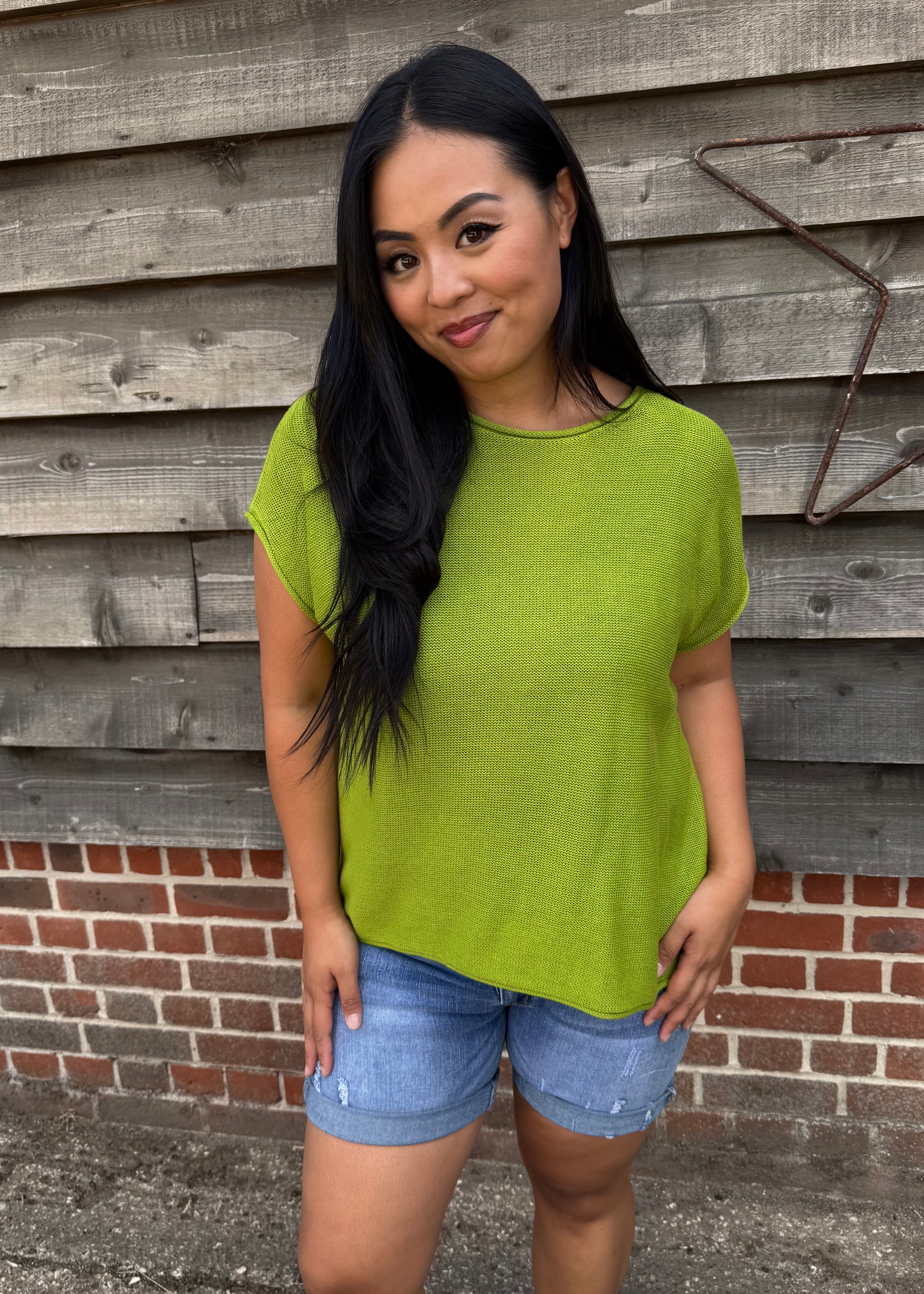 Woman wearing a green top and denim shorts standing against a wooden wall.