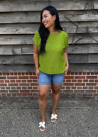 Woman wearing a green shirt and denim shorts standing against a wooden wall.