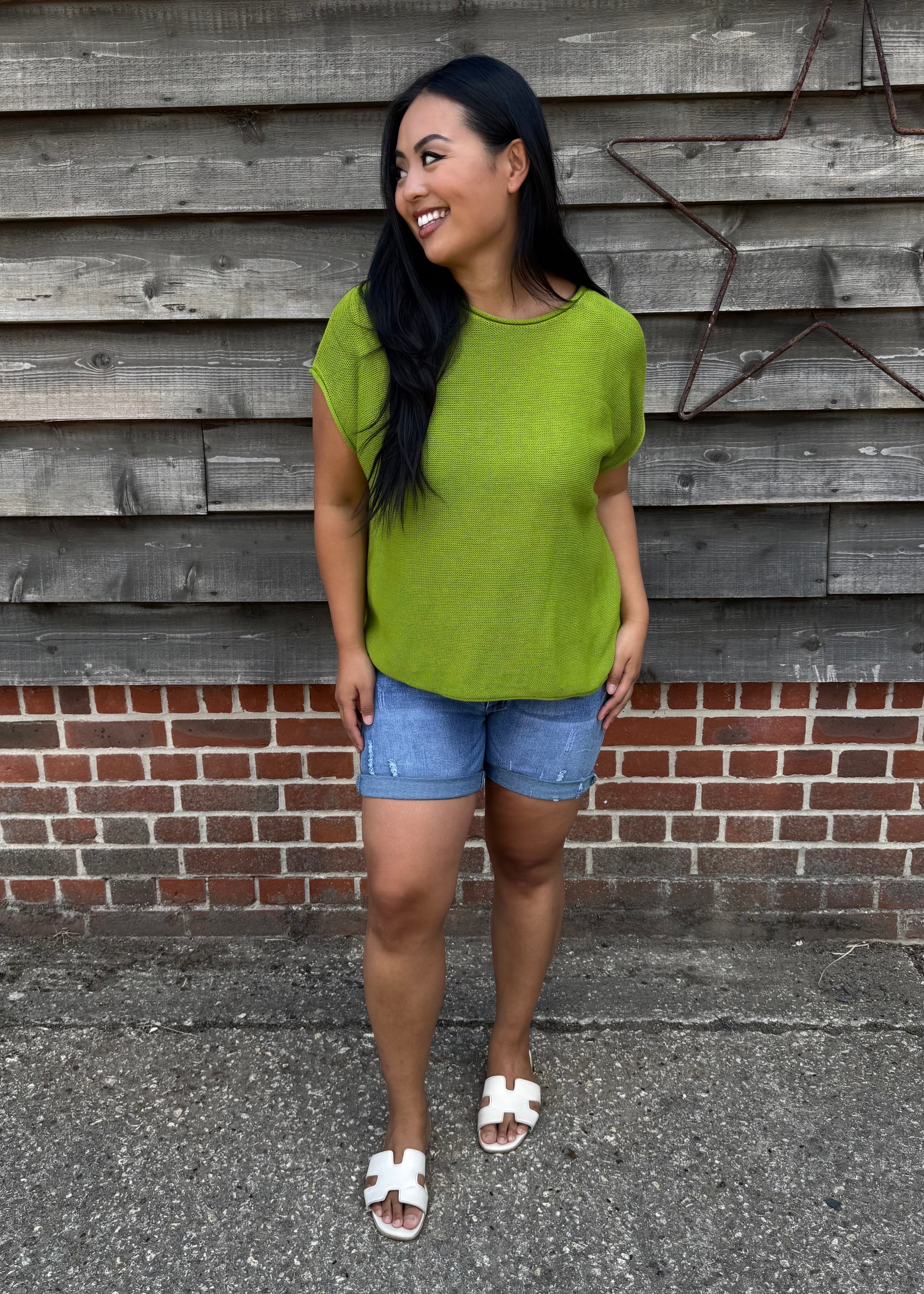 Woman wearing a green shirt and denim shorts standing against a wooden wall.