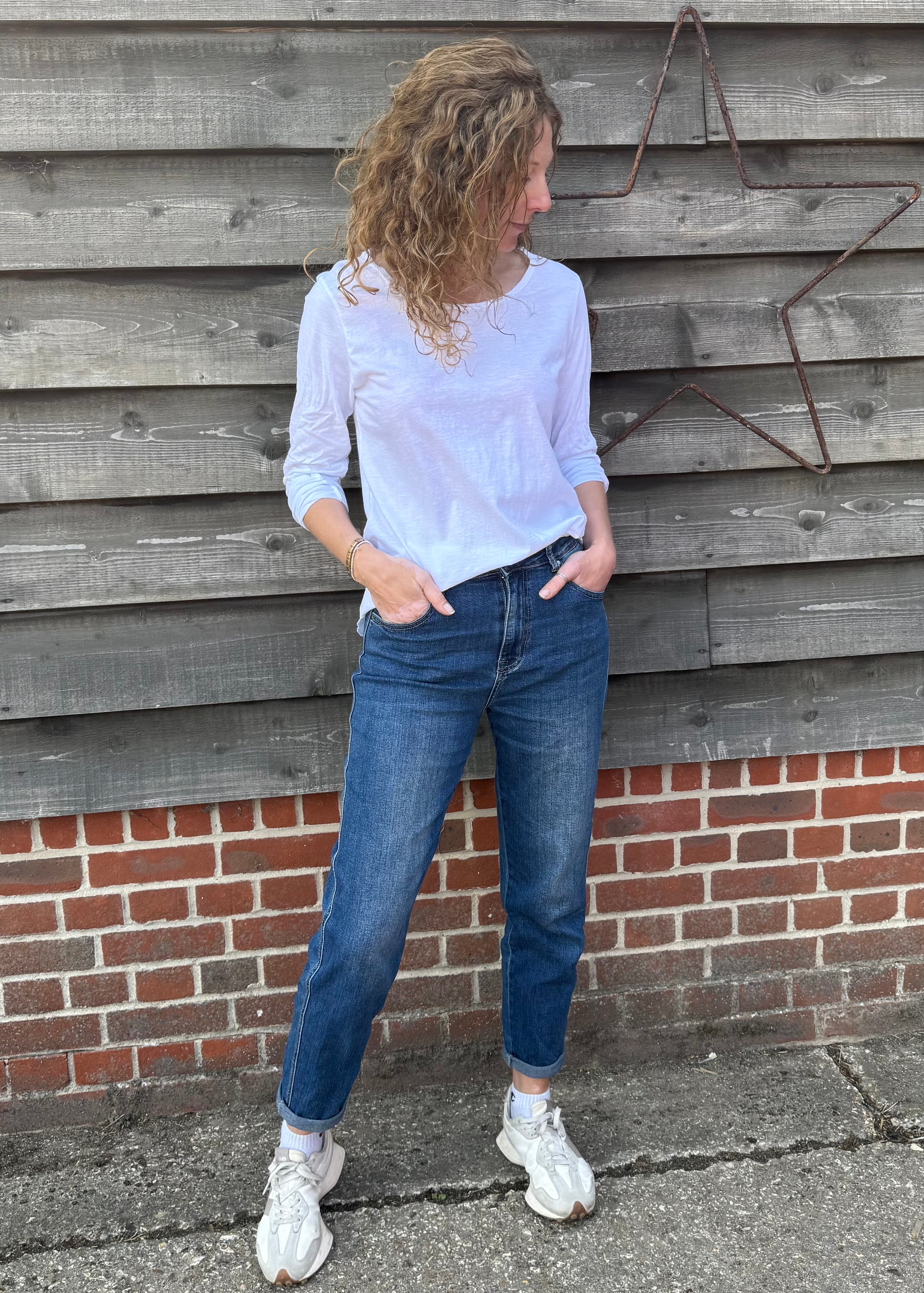 Woman wearing a white shirt and blue jeans standing against a wooden wall.