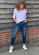 Person wearing a white shirt, blue jeans, and white sneakers leaning against a wooden wall.