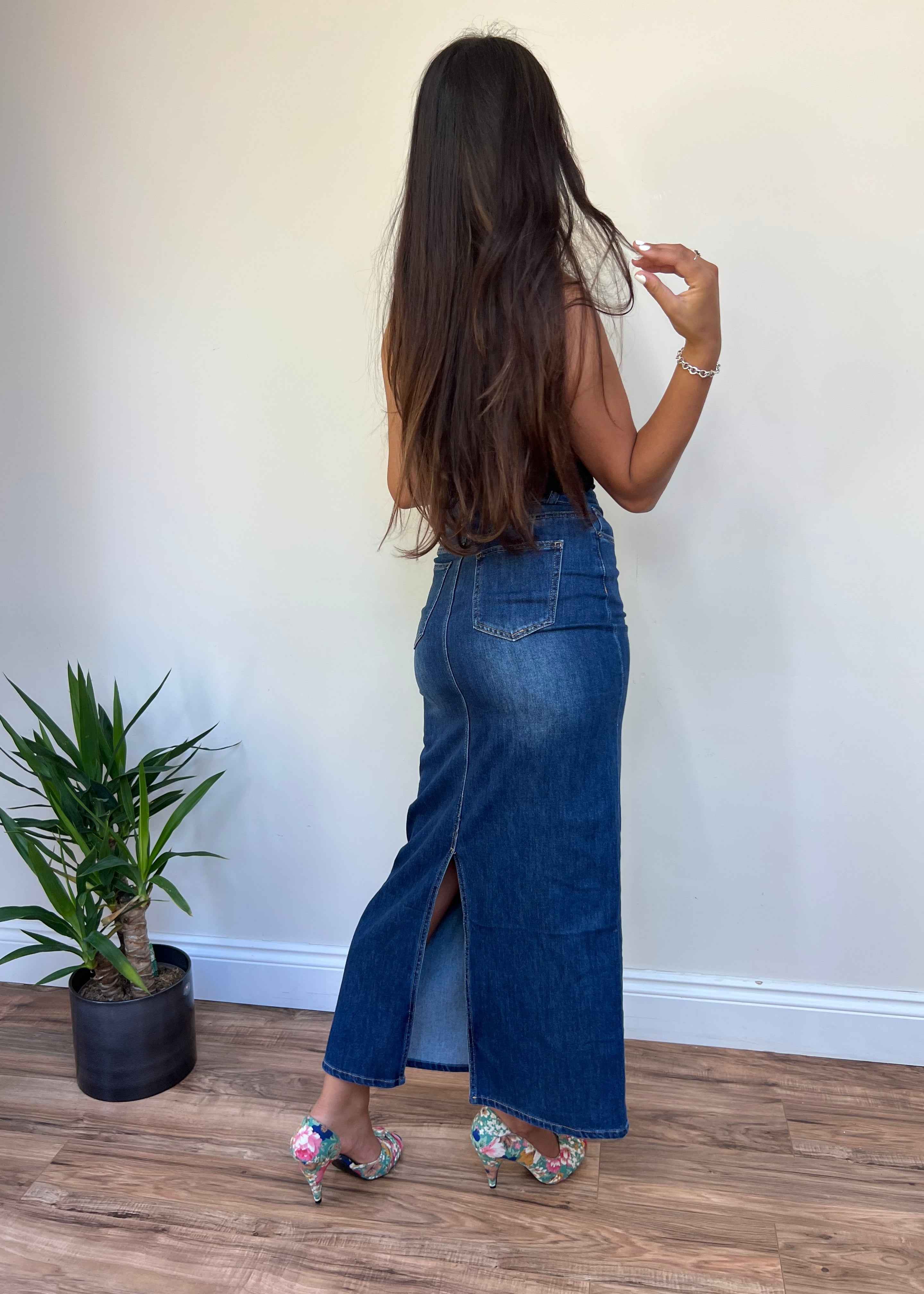 A person standing with their back to the camera wearing a denim skirt with a split at the back, paired with floral shoes and posing near a potted plant.