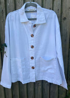 A person standing outdoors wearing a white linen mix jacket with long sleeves, blue jeans, and white shoes.