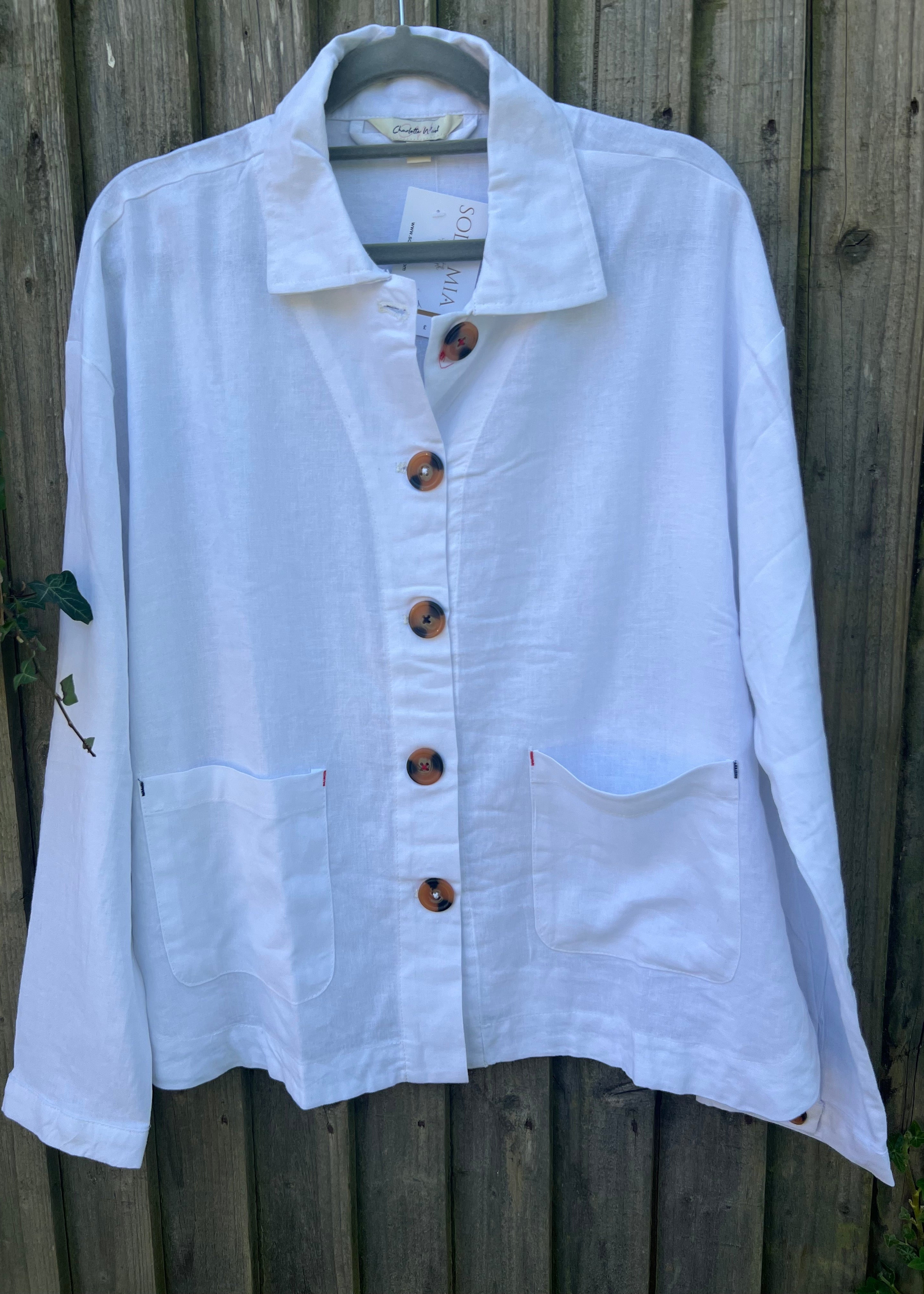 A person standing outdoors wearing a white linen mix jacket with long sleeves, blue jeans, and white shoes.