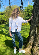 A person standing outdoors wearing a white linen mix jacket with long sleeves, blue jeans, and white shoes.