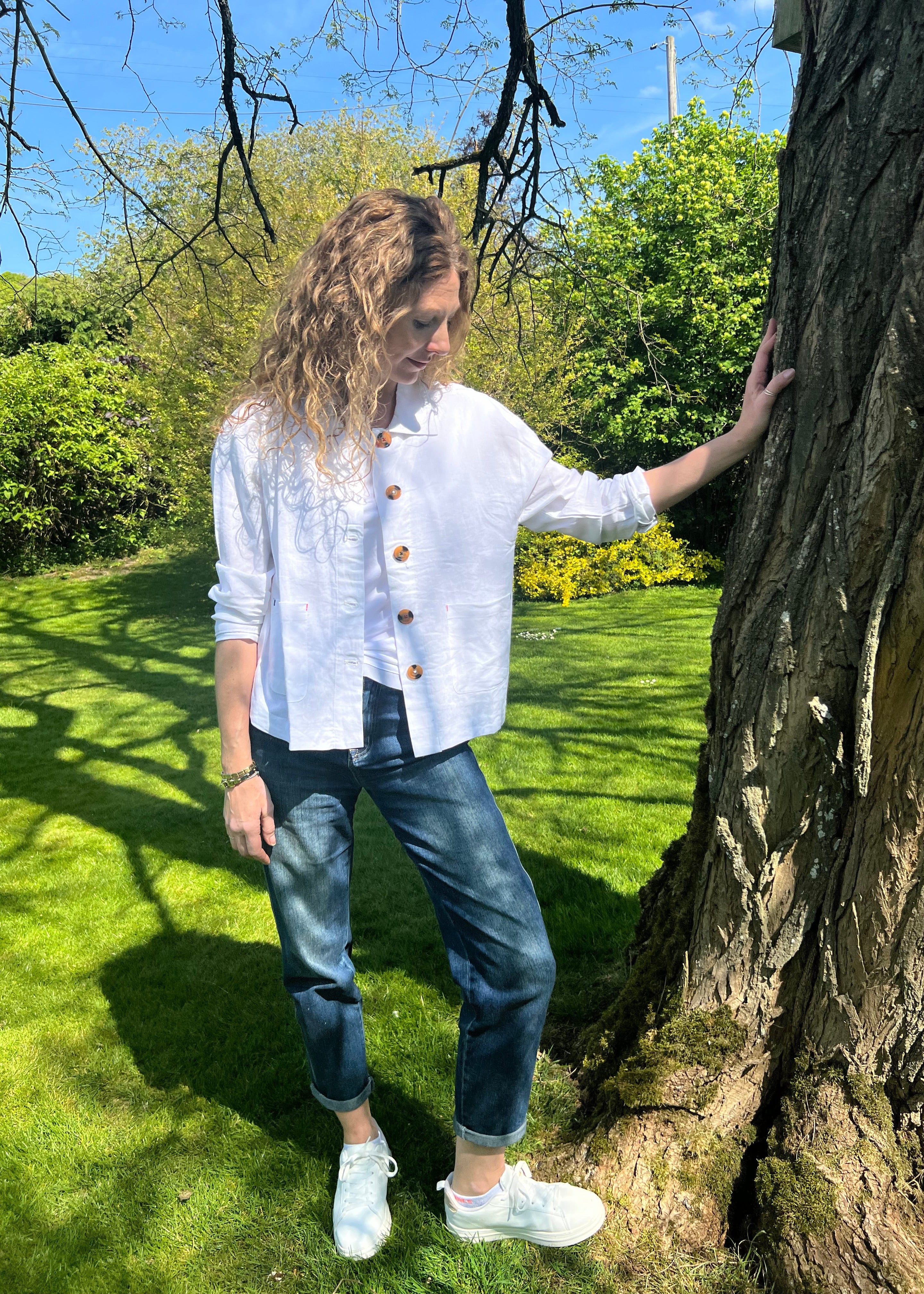 A person standing outdoors wearing a white linen mix jacket with long sleeves, blue jeans, and white shoes.