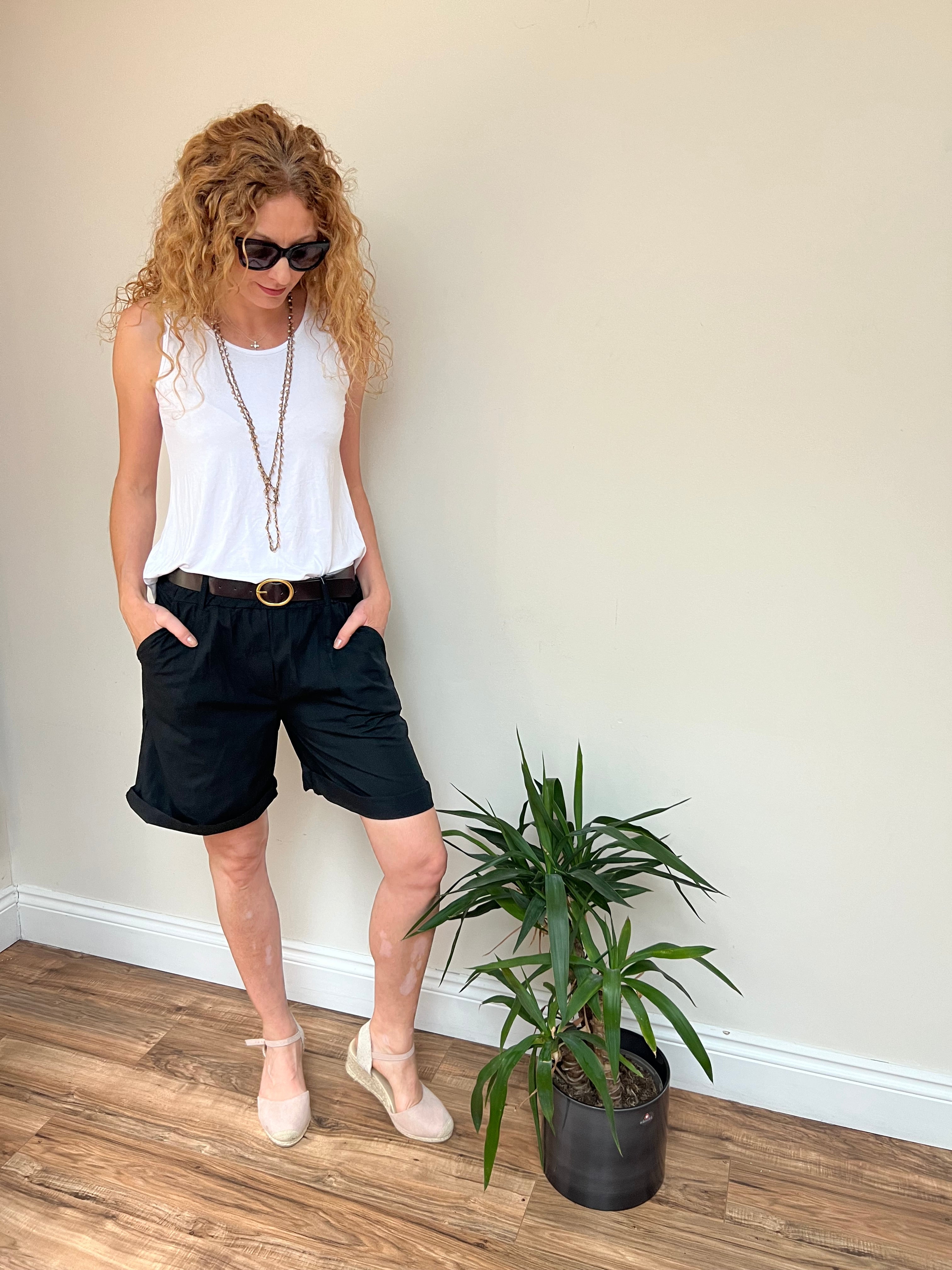 A person standing in a room with a white top and sunglasses, next to a potted plant, wearing black shorts with a brown belt.