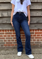 Person wearing blue jeans and a white shirt standing against a wooden and brick wall.