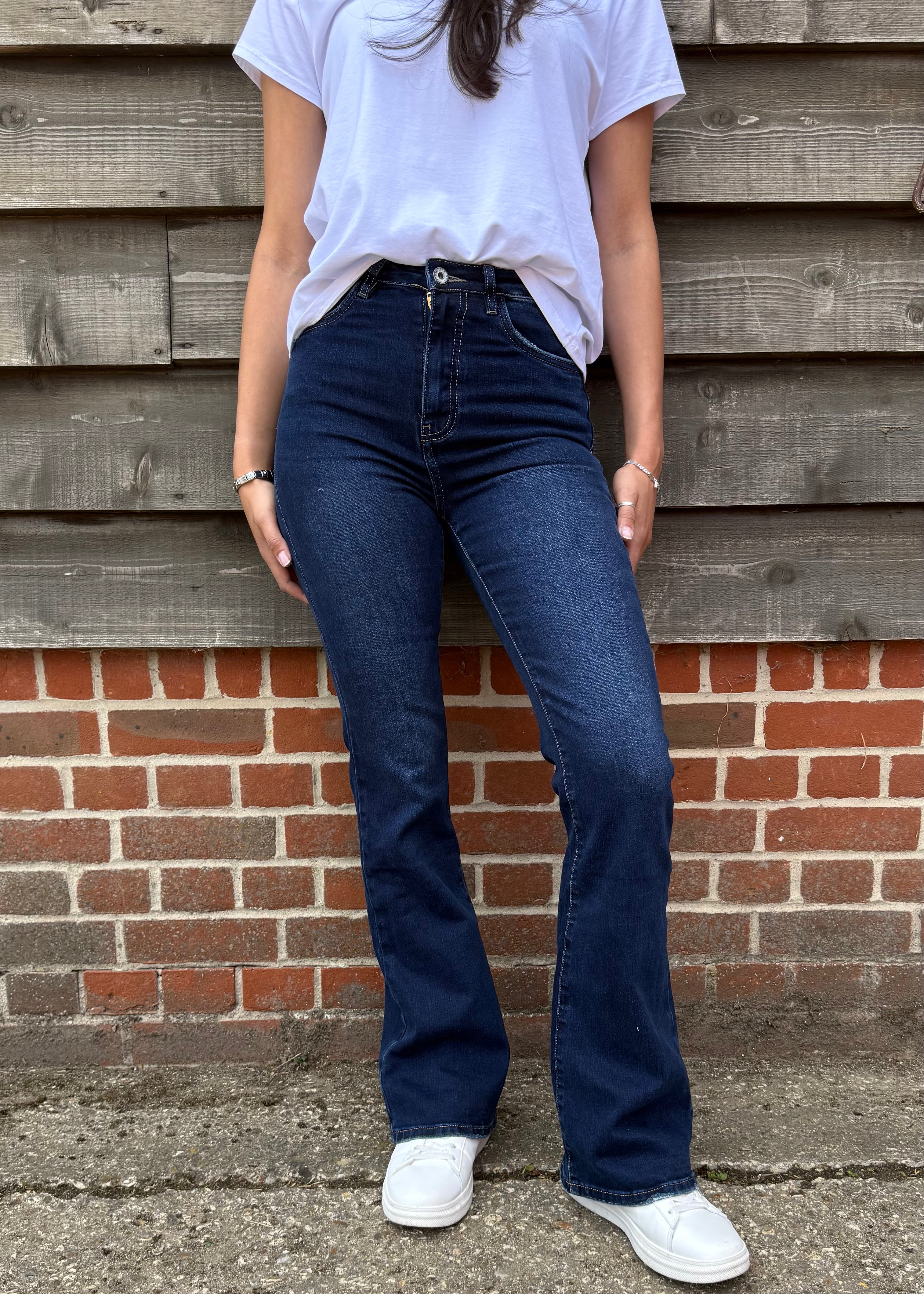 Person wearing blue jeans and a white shirt standing against a wooden and brick wall.