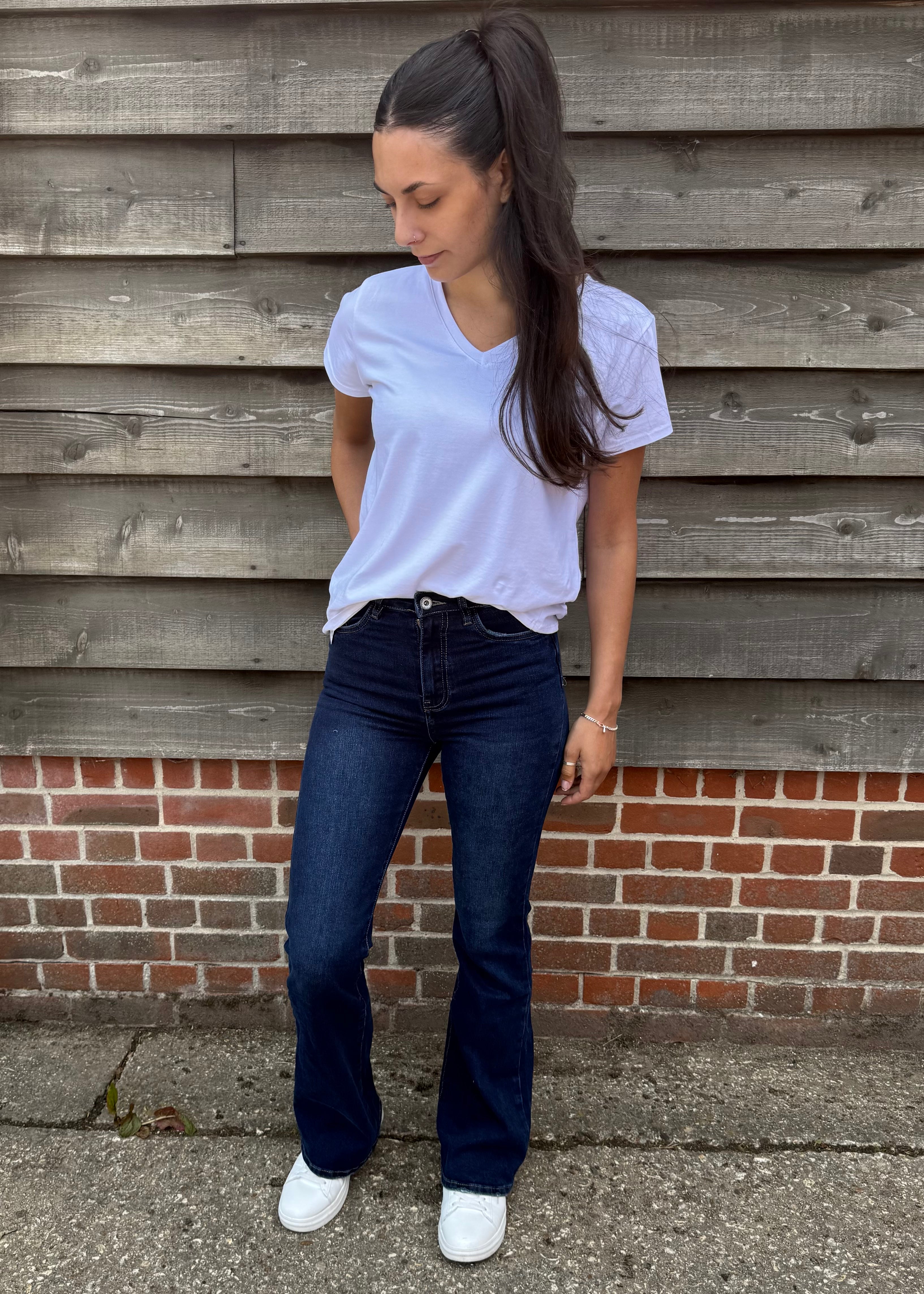 Woman wearing a white t-shirt and blue jeans standing against a wooden wall with a brick base.