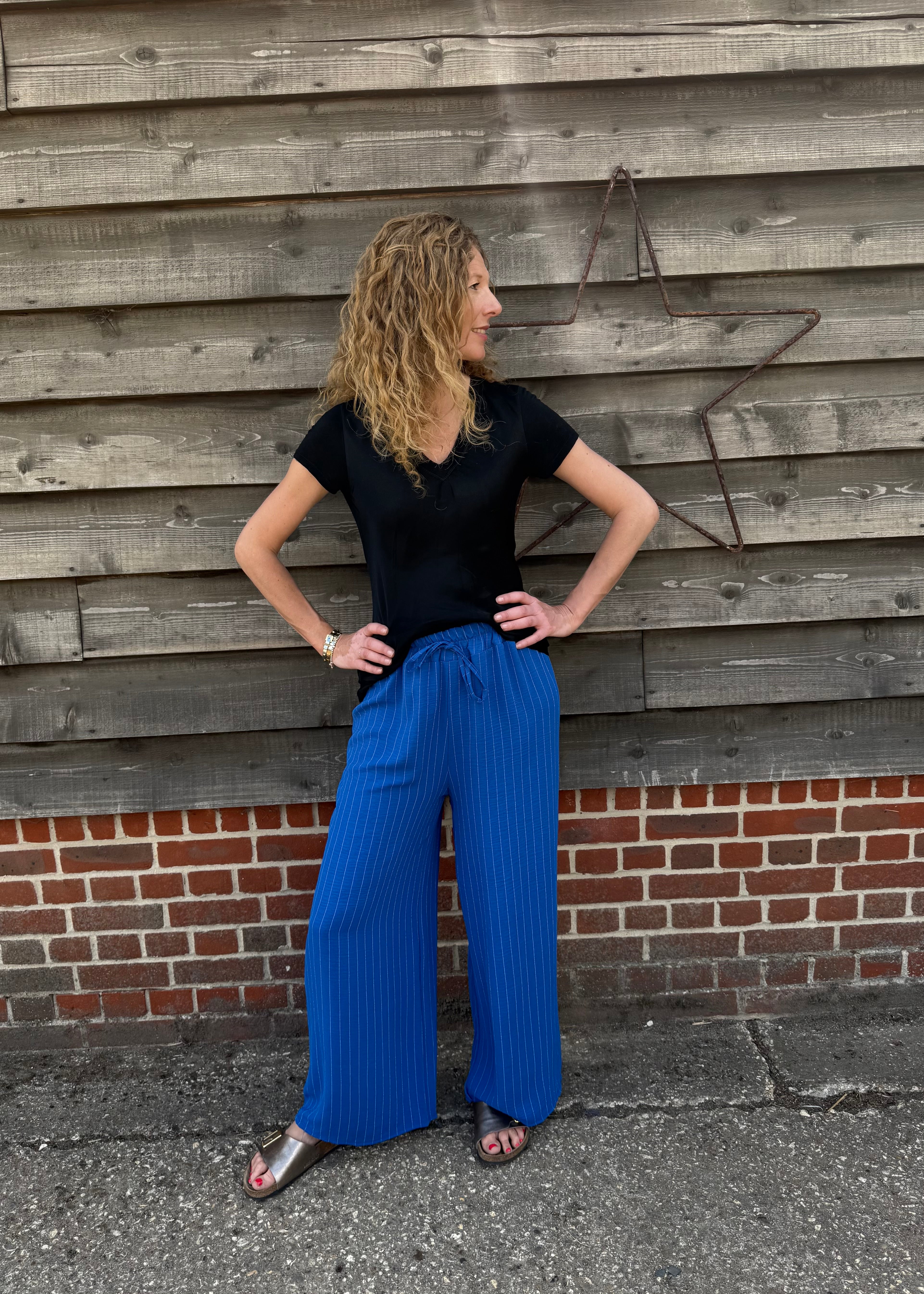 Woman wearing a black top and blue pants standing against a wooden wall.
