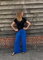 Woman wearing a black top and blue pants standing against a wooden wall.