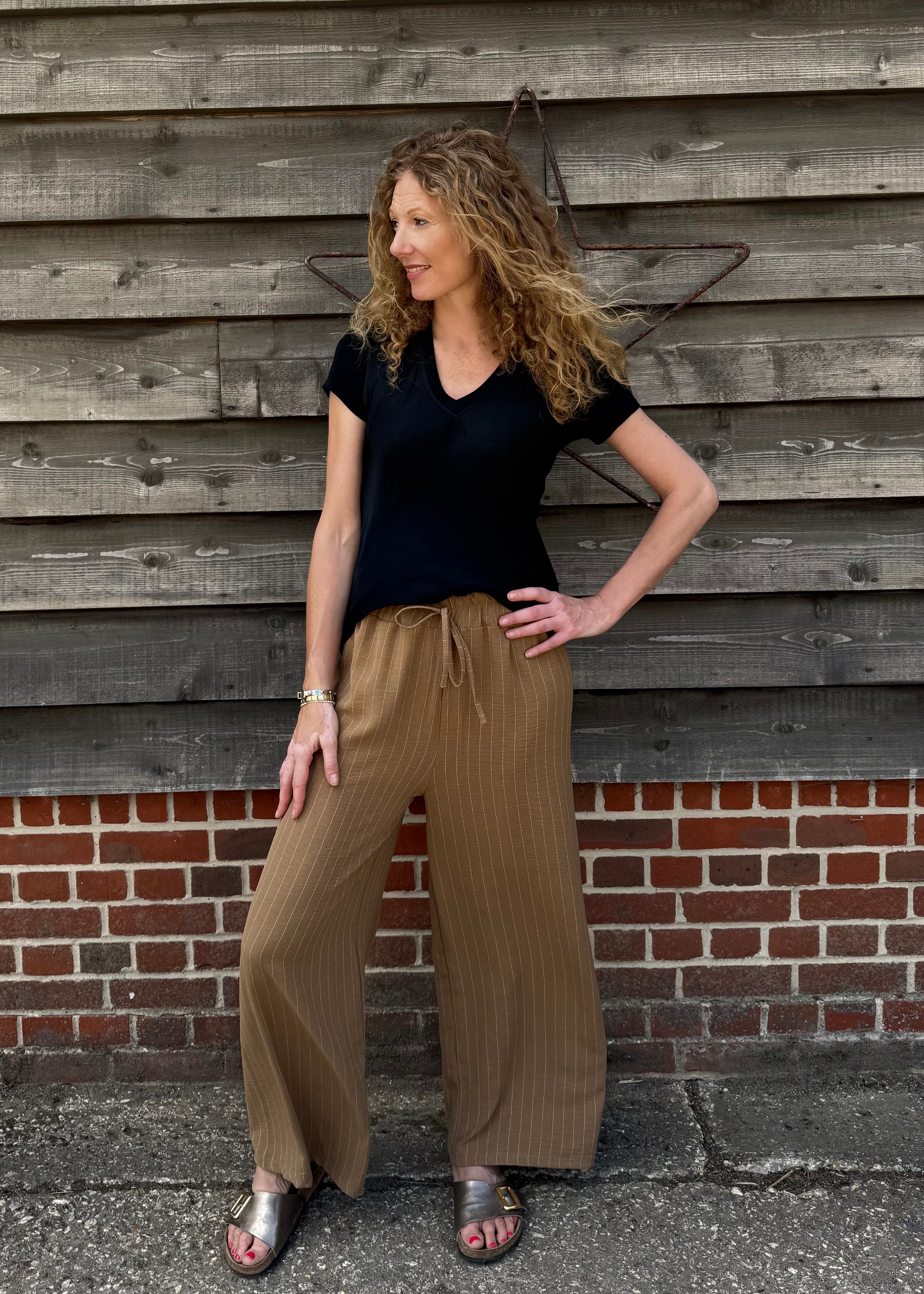 Woman wearing a black top and brown pants standing against a wooden wall.