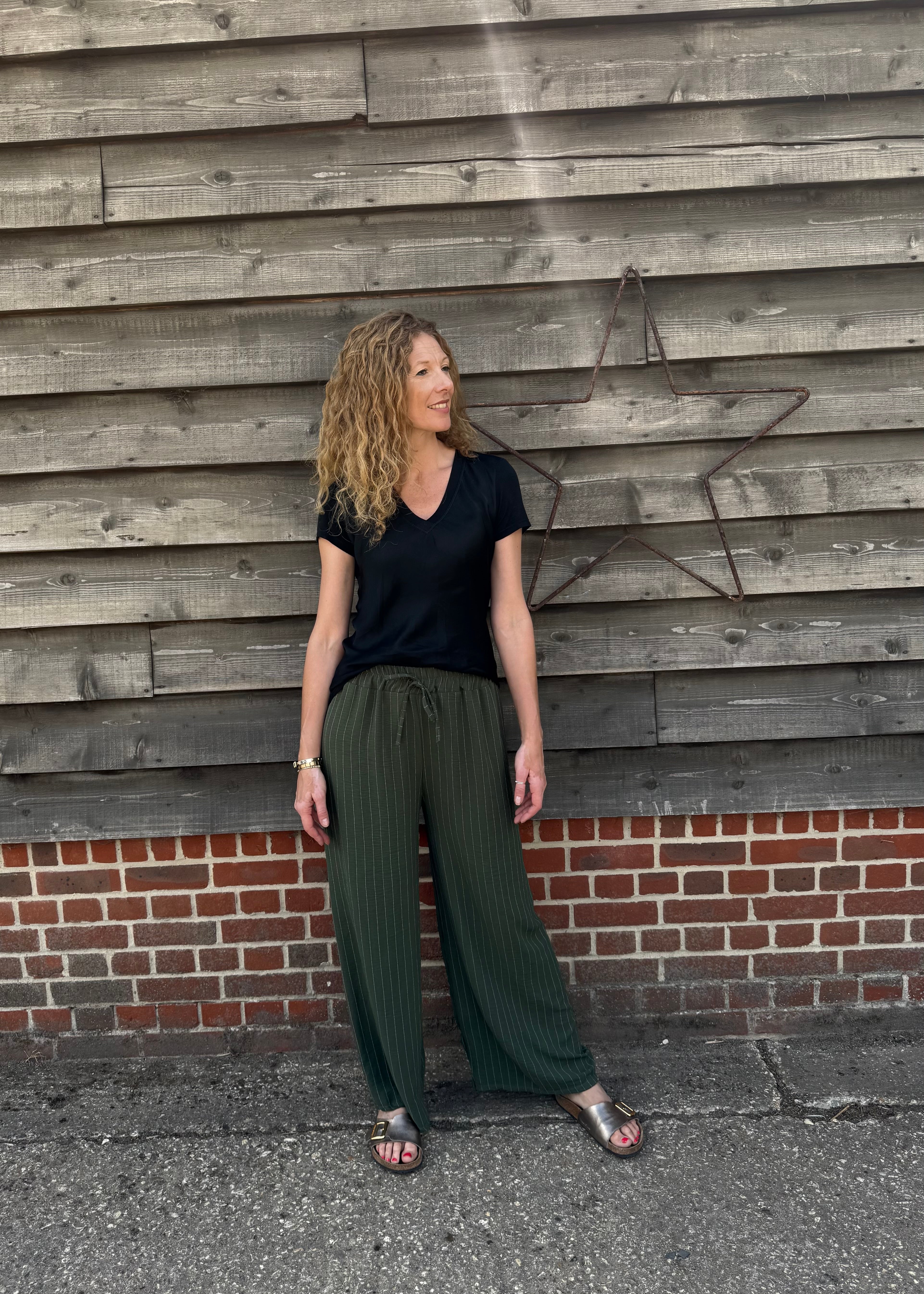 Woman wearing a black top and green pants standing against a wooden wall.