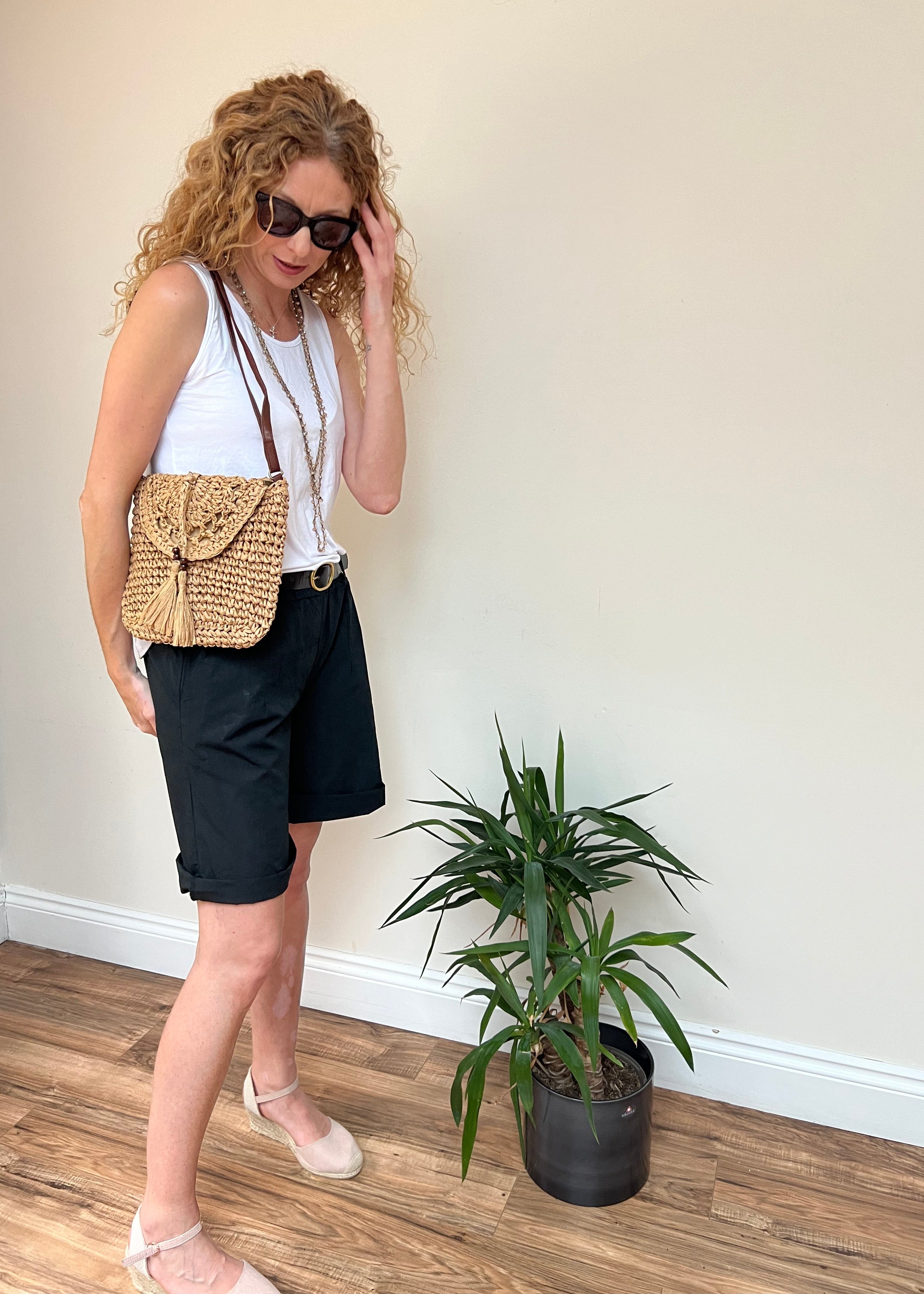 A woman wearing sunglasses and a white top with a brown belt, carrying a brown straw bag, standing next to a potted plant.