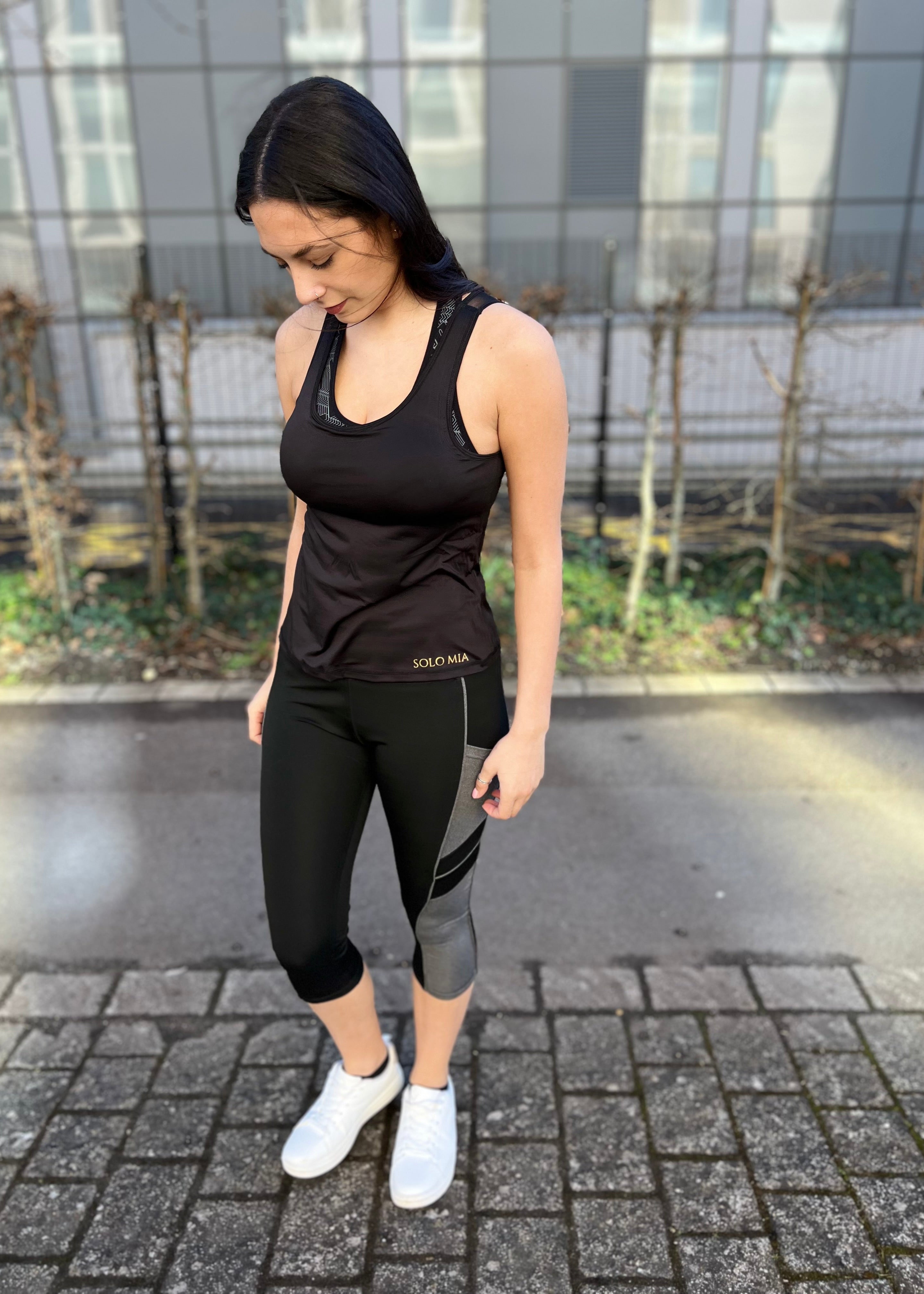 A person wearing black 3/4 length leggings with side pockets, paired with a sleeveless top and white sneakers, standing on a brick pathway.