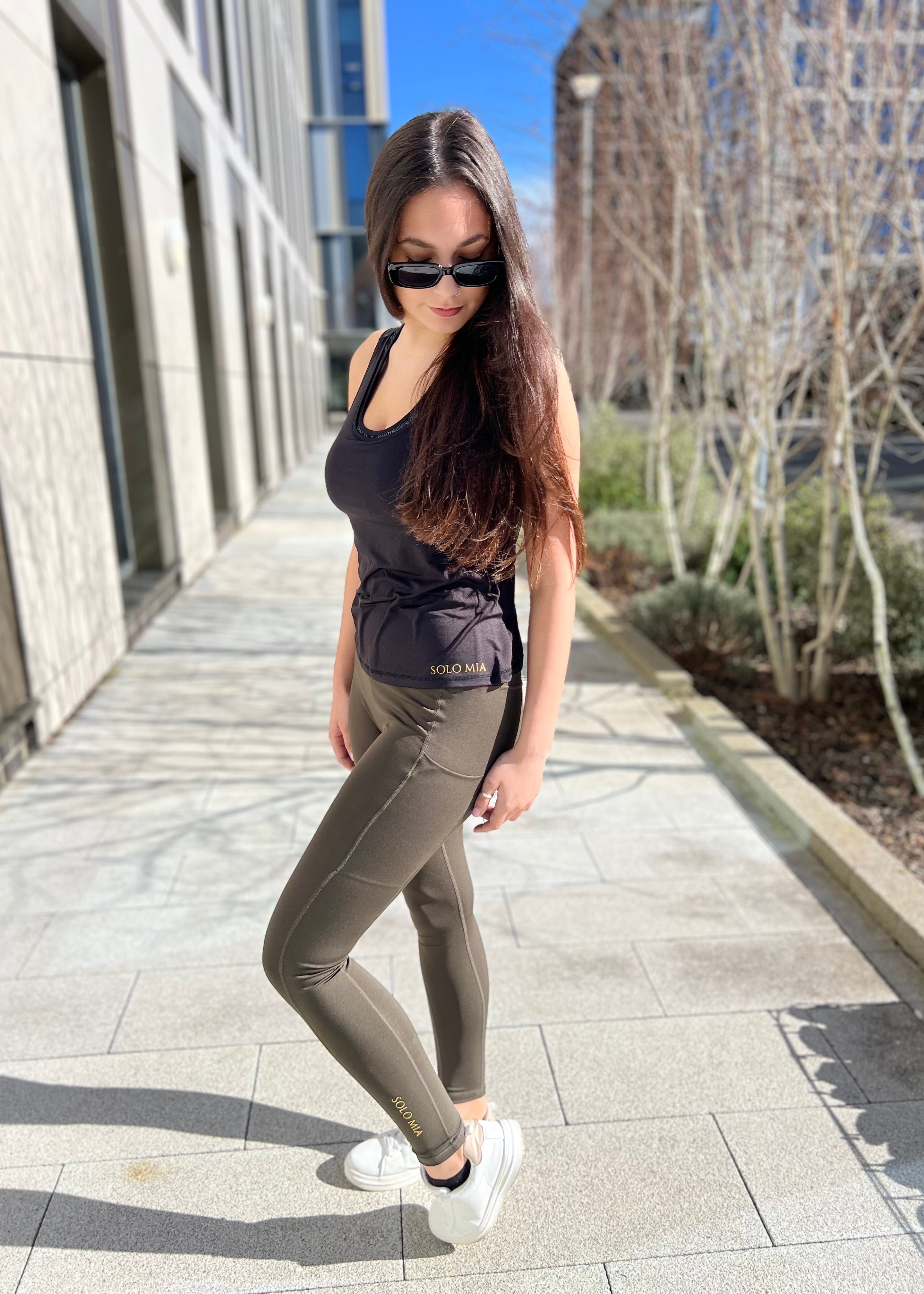 A woman standing on a sidewalk wearing sportiva running leggings, a tank top, sunglasses, and white sneakers.