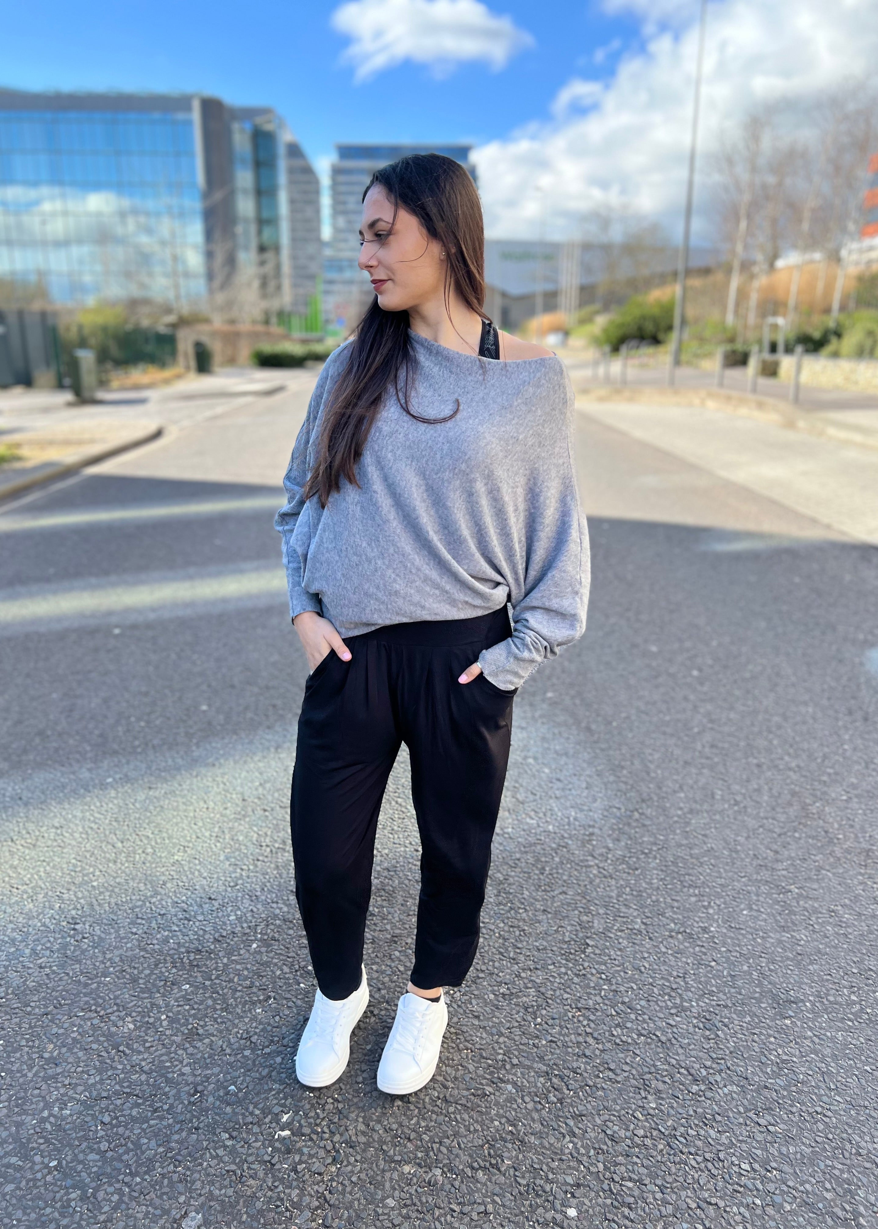 A woman standing on the street wearing black slouch joggers and white sneakers, with a grey sky and buildings in the background.