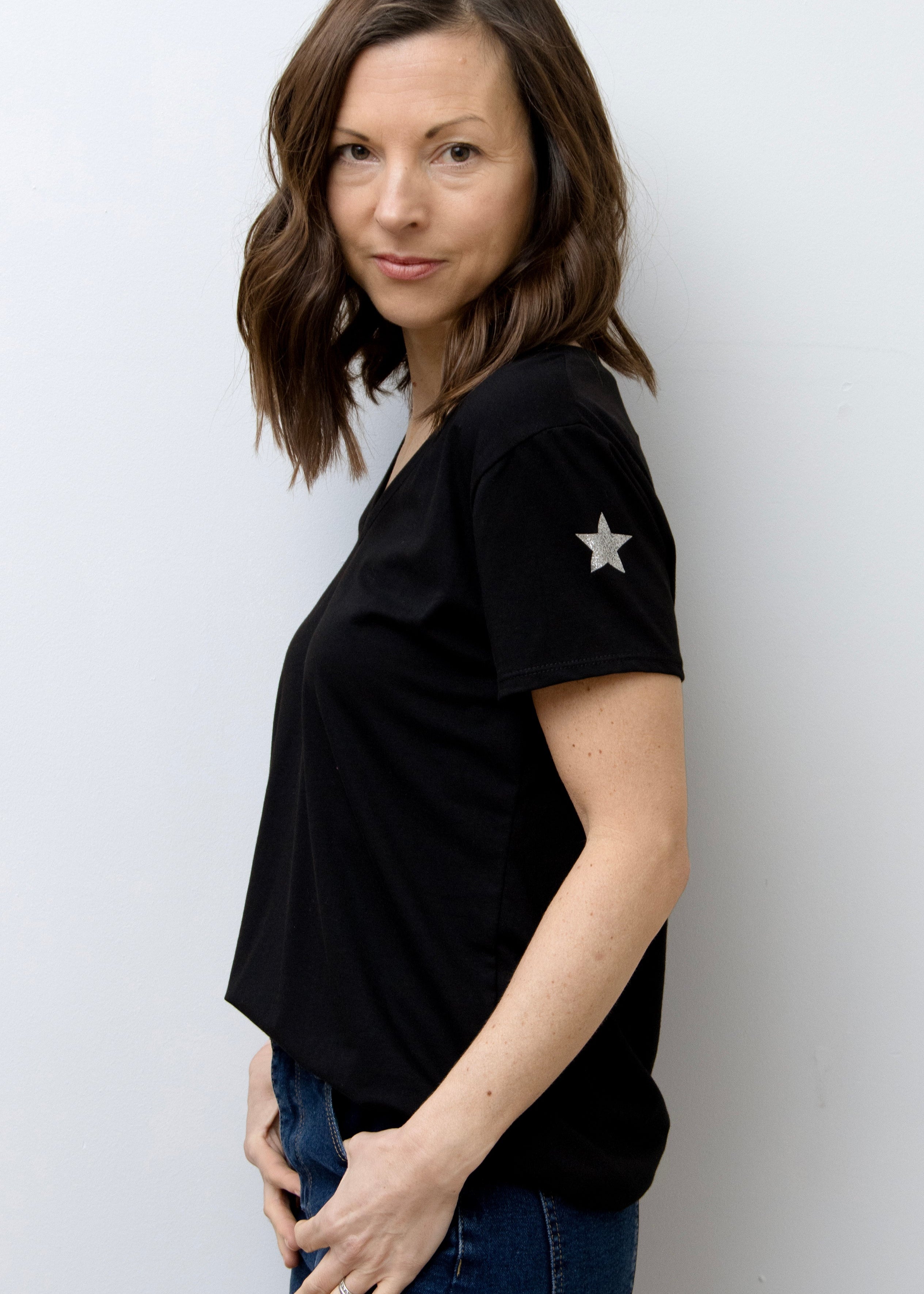 A woman smiling, wearing a pink V-neck T-shirt with a silver star on the sleeve, paired with blue jeans.