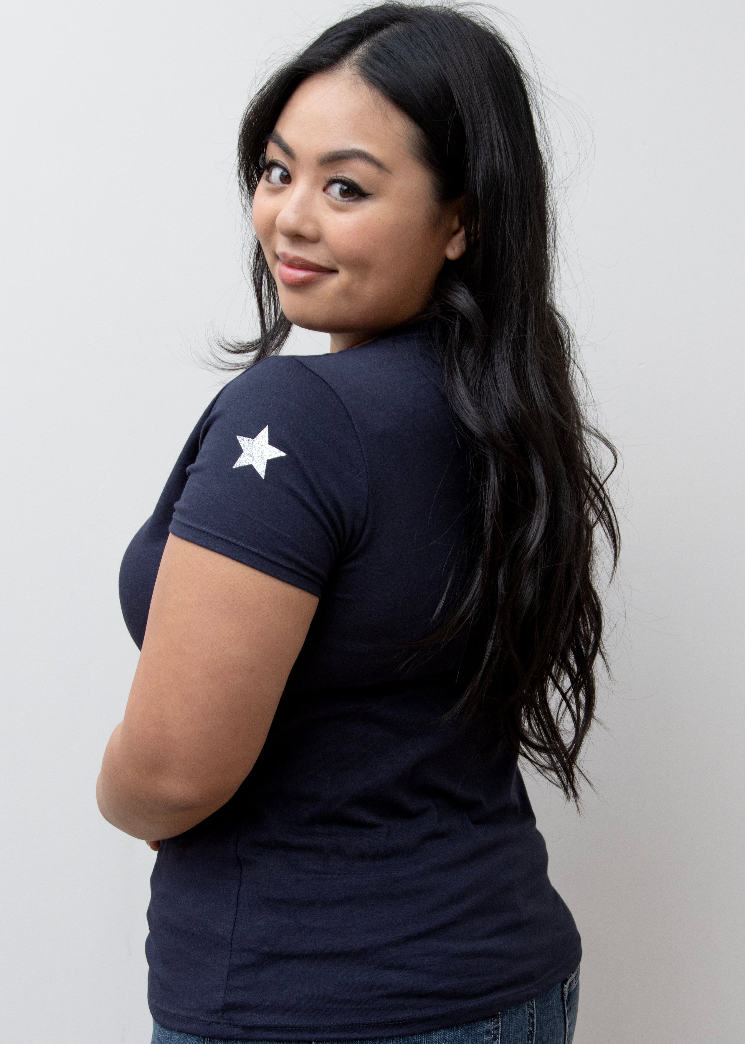 A woman smiling, wearing a pink V-neck T-shirt with a silver star on the sleeve, paired with blue jeans.