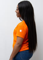 A woman smiling, wearing a pink V-neck T-shirt with a silver star on the sleeve, paired with blue jeans.