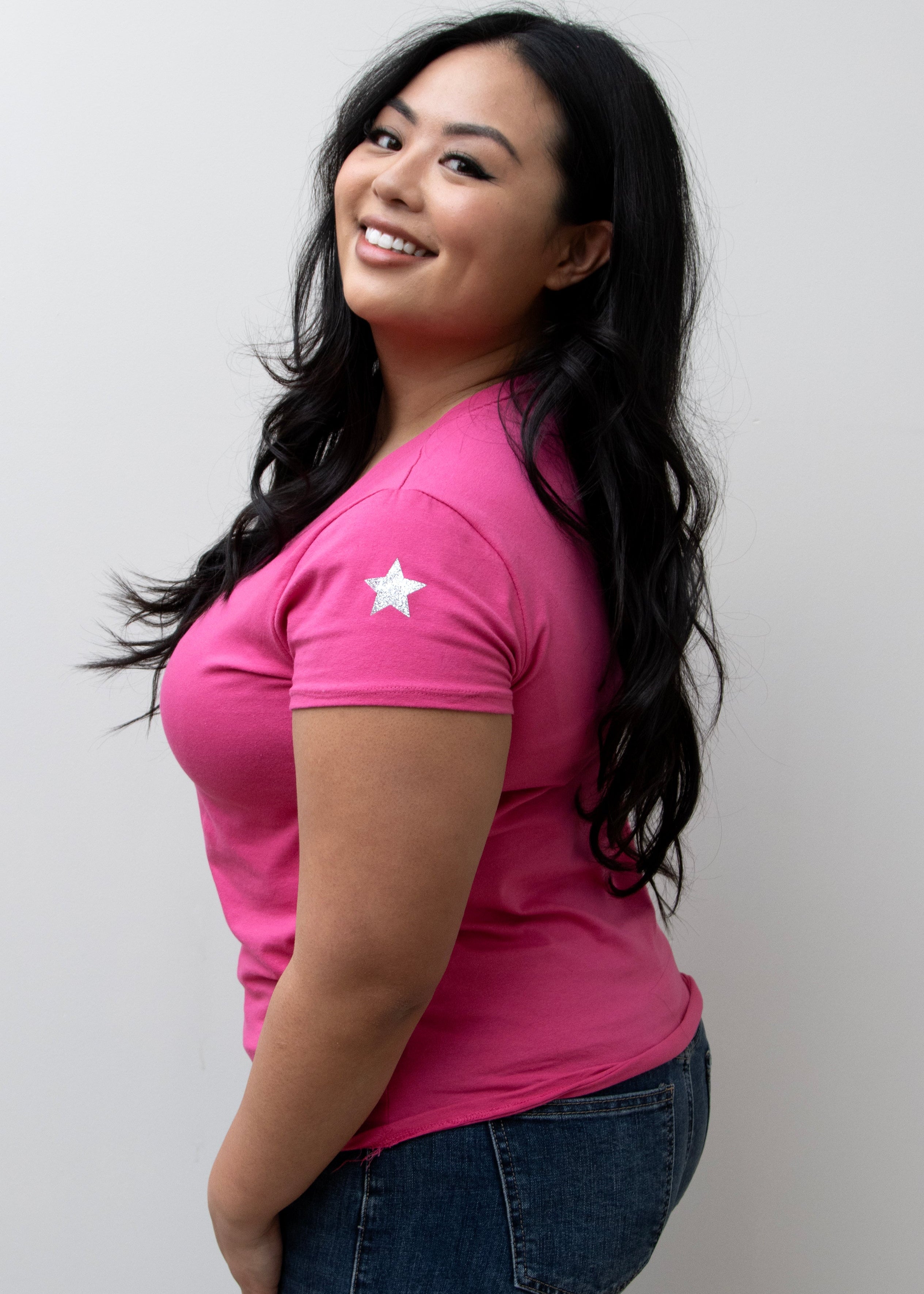 A woman smiling, wearing a pink V-neck T-shirt with a silver star on the sleeve, paired with blue jeans.