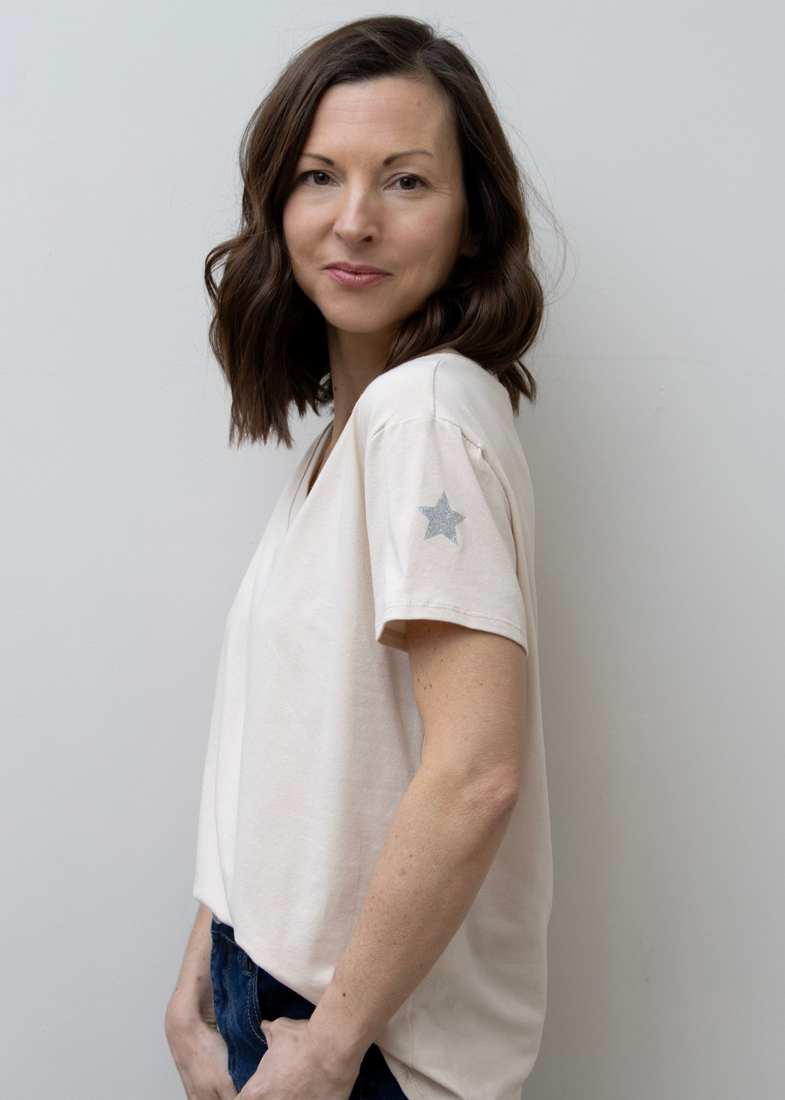 A woman smiling, wearing a pink V-neck T-shirt with a silver star on the sleeve, paired with blue jeans.