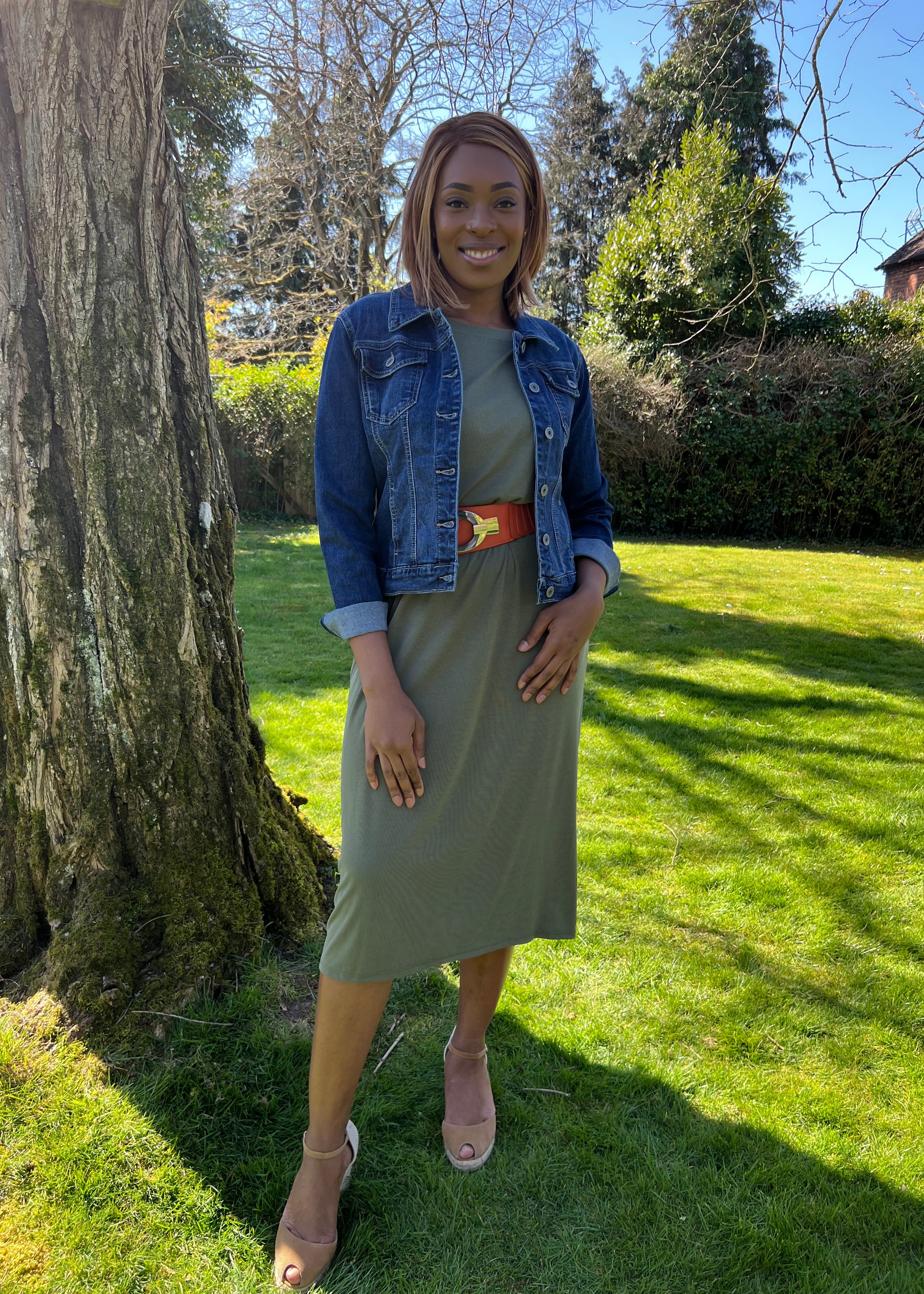 A woman standing outdoors wearing a green midi dress with a round neckline, sleeveless design, and knee-length. She has on a blue denim jacket, beige belt, and matching beige shoes.