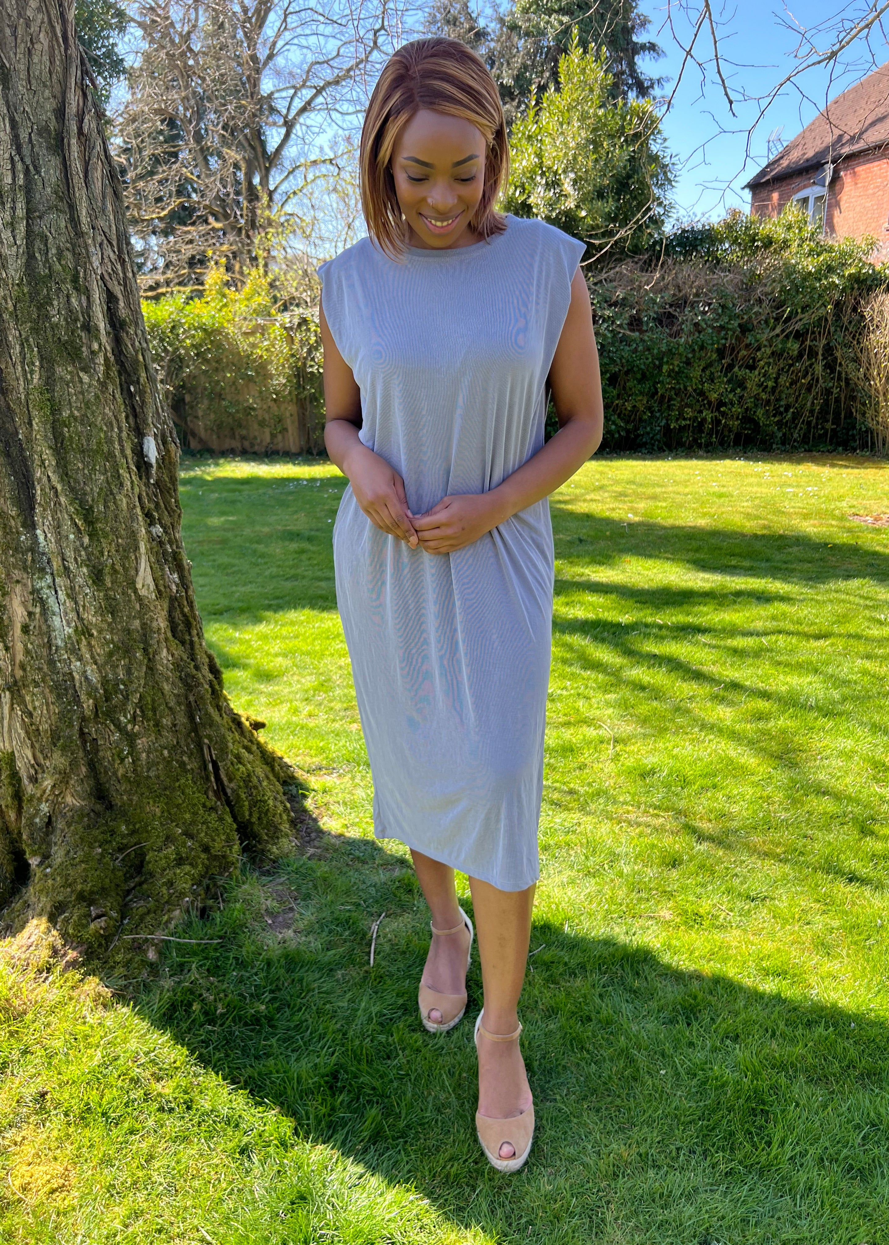 A woman standing outdoors wearing a green midi dress with a round neckline, sleeveless design, and knee-length. She has on a blue denim jacket, beige belt, and matching beige shoes.