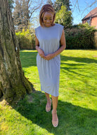 A woman standing outdoors wearing a green midi dress with a round neckline, sleeveless design, and knee-length. She has on a blue denim jacket, beige belt, and matching beige shoes.