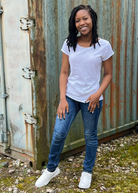 A woman standing in front of a corrugated metal wall, wearing a white t-shirt, blue jeans, and white sneakers.
