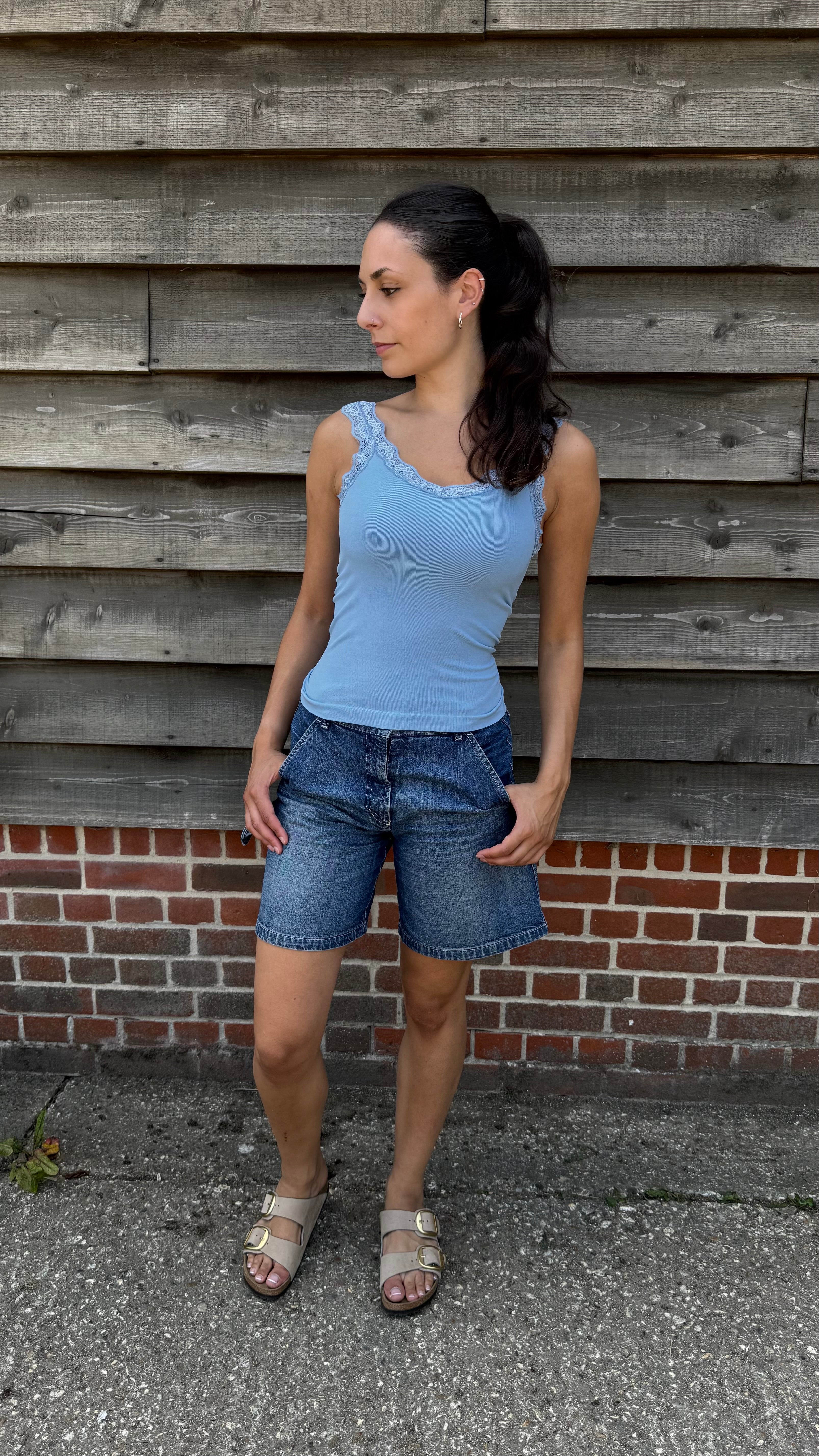 Woman wearing a blue tank top and denim shorts standing against a wooden wall.