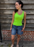 Woman wearing a bright green tank top and blue shorts standing against a wooden wall.