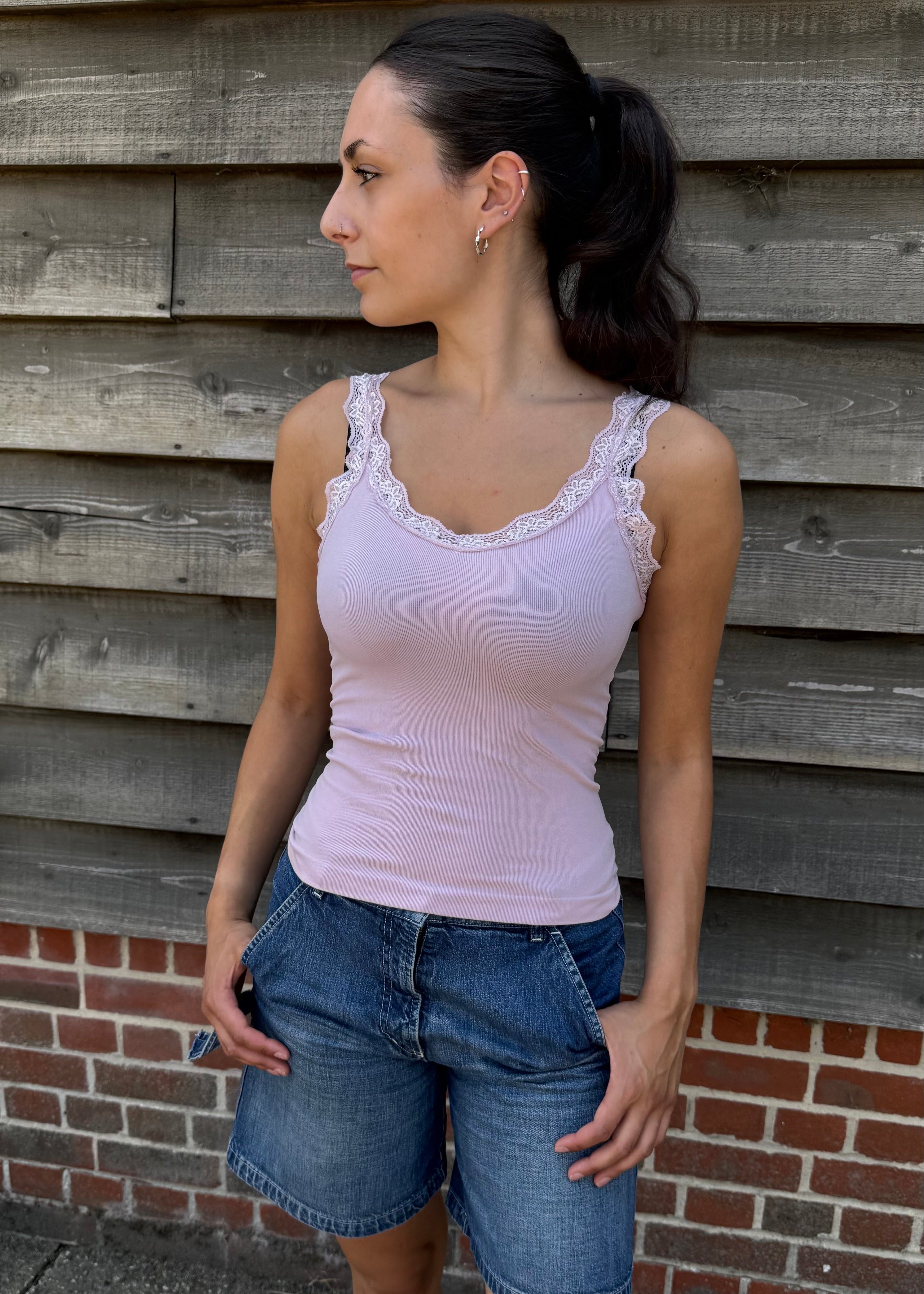 Woman wearing a light purple tank top and denim shorts standing against a wooden wall.