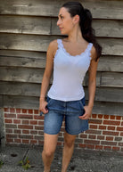 Woman wearing a white tank top and denim shorts standing against a wooden wall.