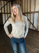 A woman standing in a room with wooden walls, wearing a long-sleeved, striped, sparkly top and blue jeans.